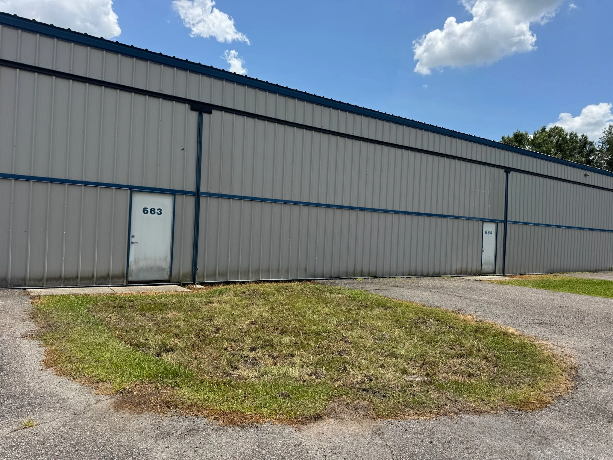 A metal building with tan walls and a blue roof, numbered 663 and 664, situated next to a sidewalk and a grassy area under a partly cloudy sky. Sod restoration.