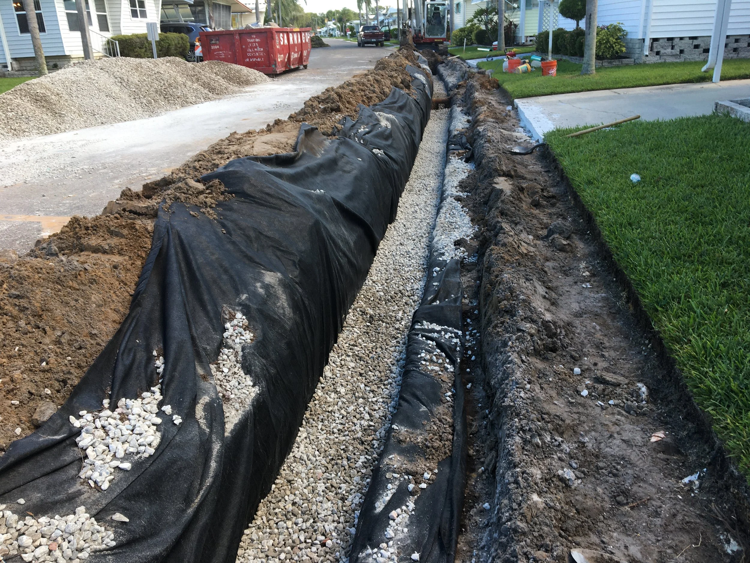 Construction site with a trench, black plastic lining, gravel, and dirt, surrounded by residential houses and construction equipment. installation of an underdrain system.