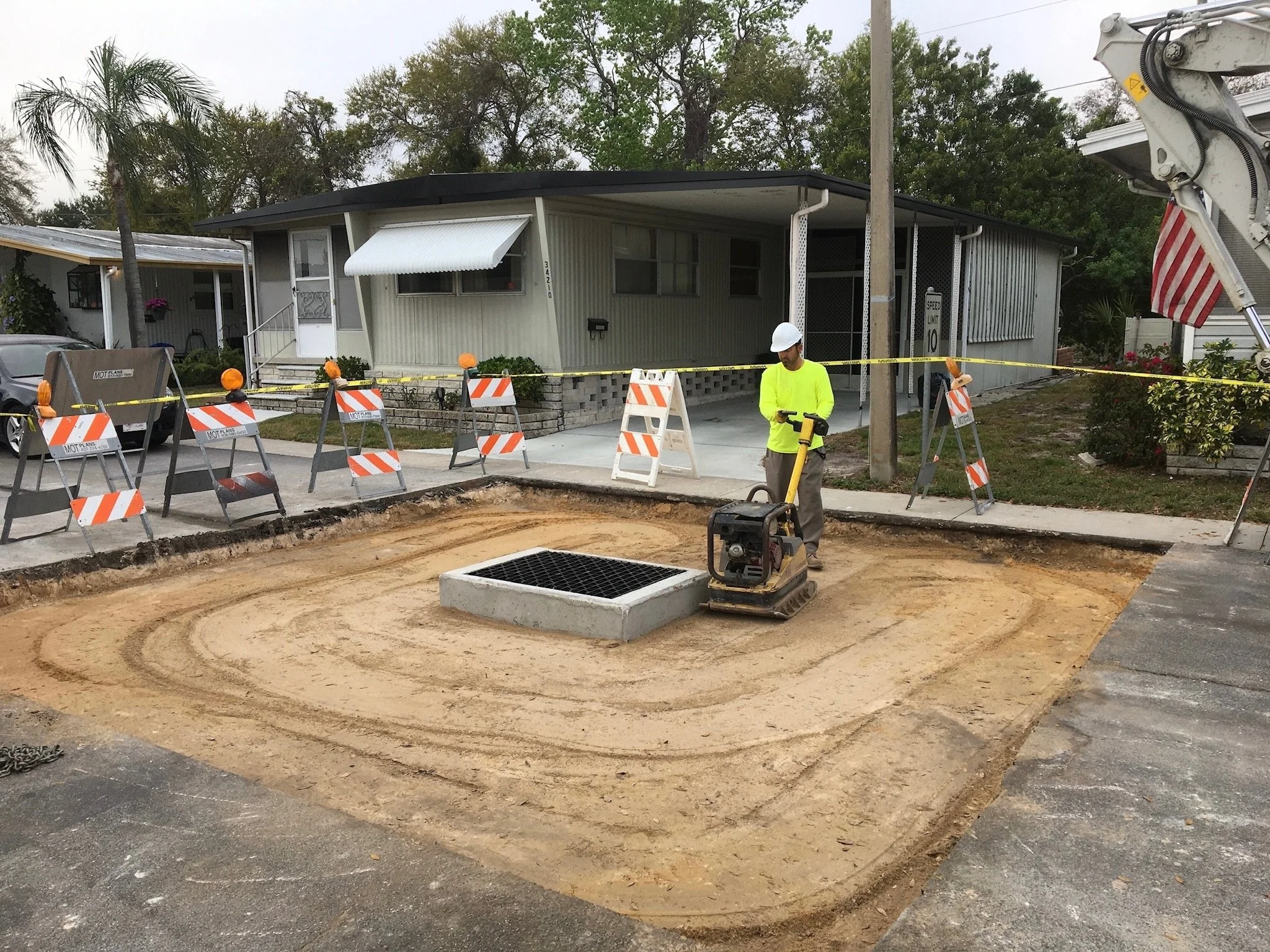 Construction site in front of a house with a worker and safety barriers, and a dirt area being prepared for paving. Compaction around structure replacement.
