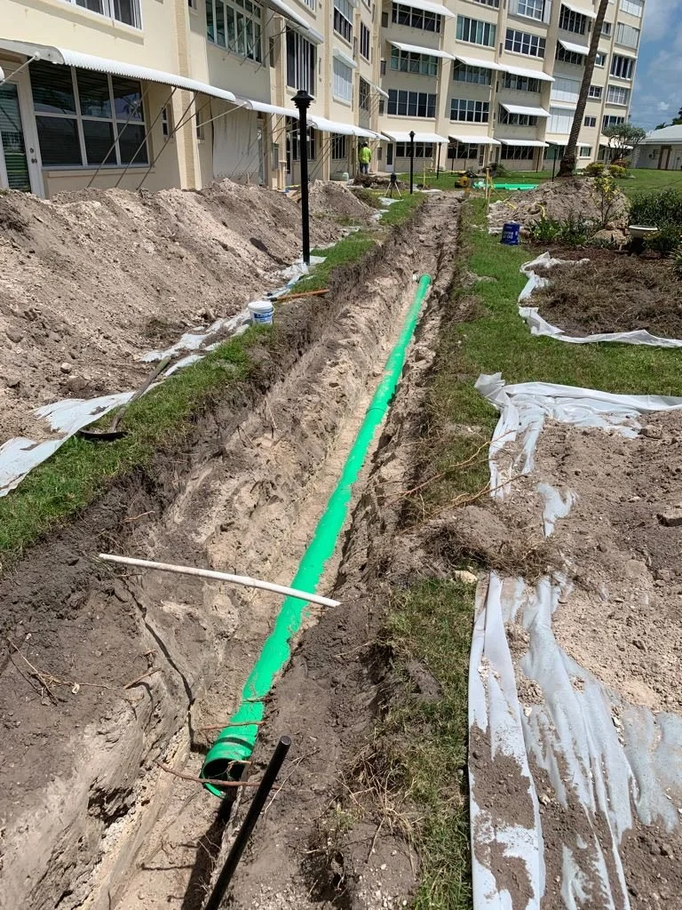 An infrastructure construction site with a long trench in the ground containing a large green pipe, surrounded by soil piles and construction materials, with an apartment building in the background.