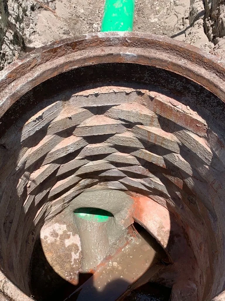 Inside a brick storm manhole with water flowing at the bottom. A green pipe outlet is visible at the top, releasing water into the pipe. Sunlight casts shadows inside the pipe.