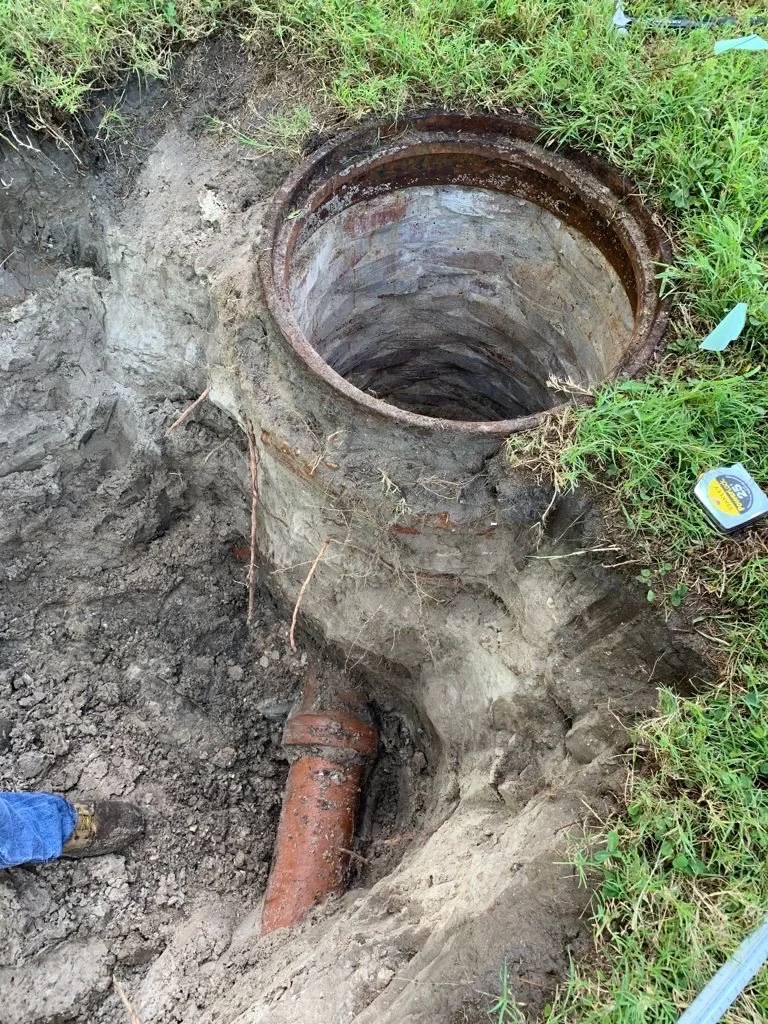 An open manhole with a pipe extending from the ground near the edge, surrounded by grass and dirt. Old clay pipe removal.
