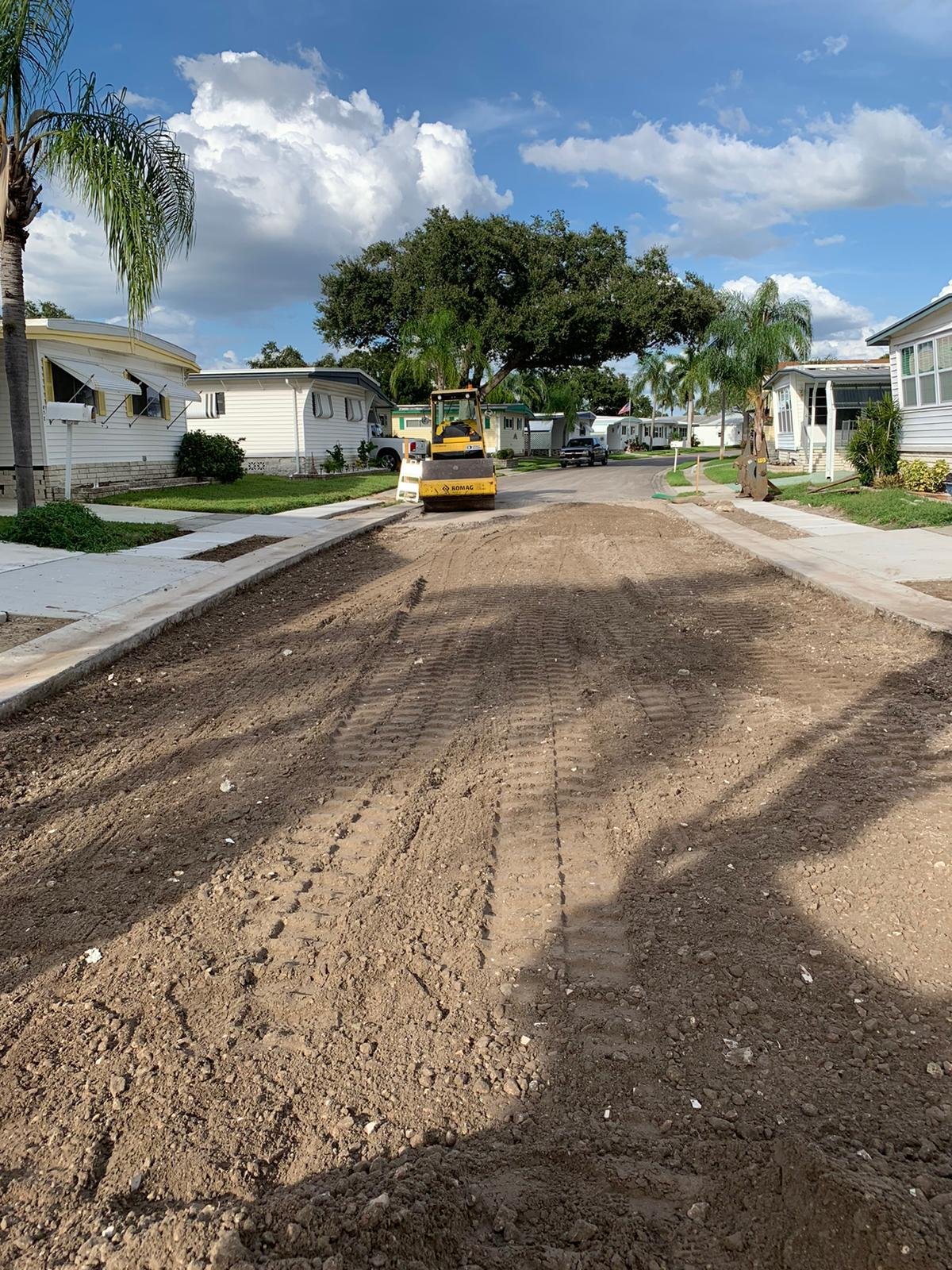 A residential street under construction with a roller compactor in the center, dirt and tire tracks in the foreground, and houses, trees, and parked cars in the background under a partly cloudy blue sky. 