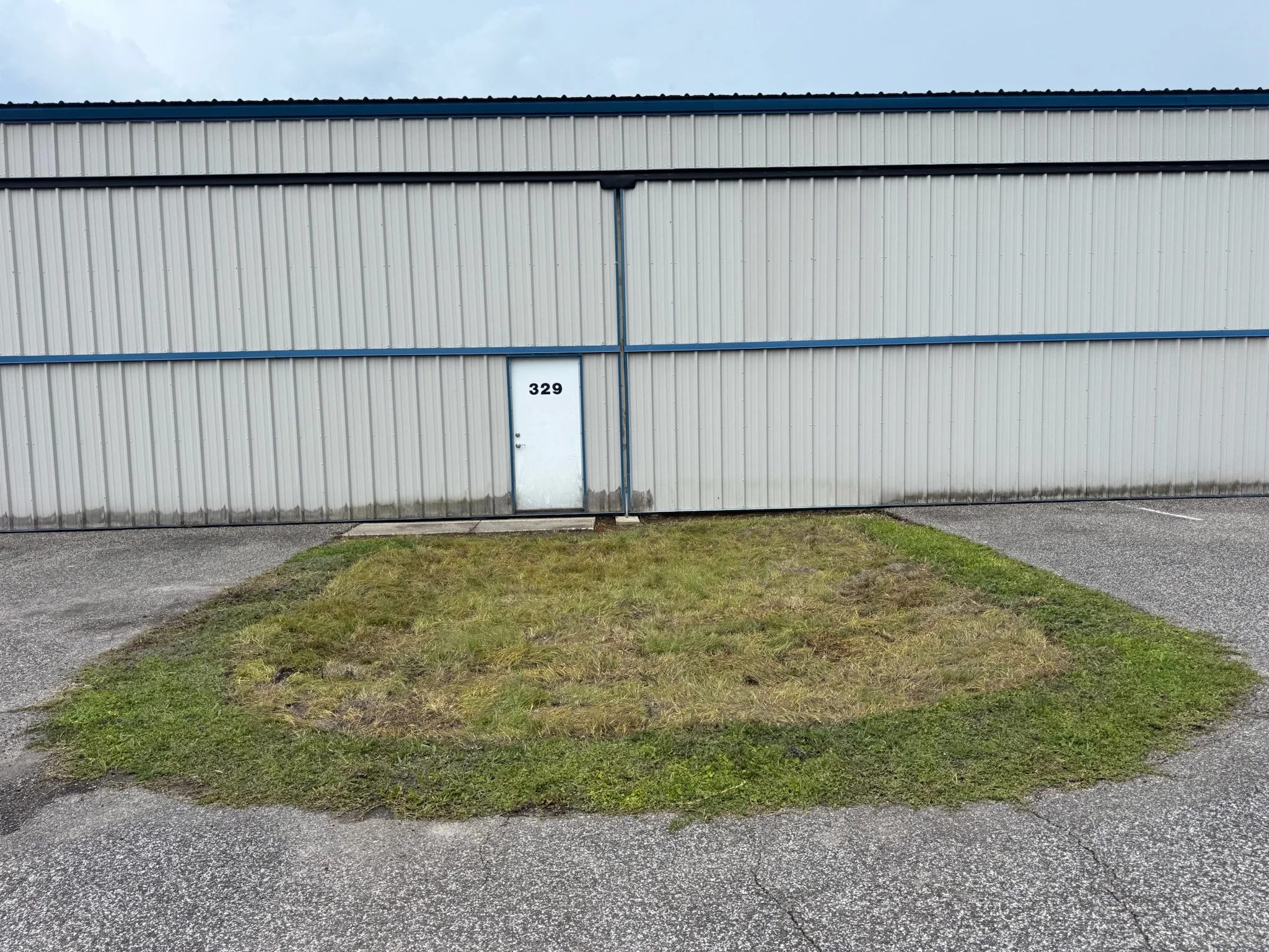 A metal building with a white corrugated exterior and a blue roof, featuring a door with the number 329, situated on a small patch of grass with a concrete walkway, under a cloudy sky.  Sod restoration.
