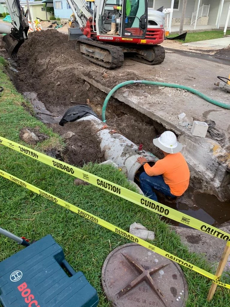 Construction worker in orange shirt and white hard hat working on large pipe in trench. Excavator parked on street, yellow caution tape surrounds work area, green hose runs through trench, tools and equipment nearby.
