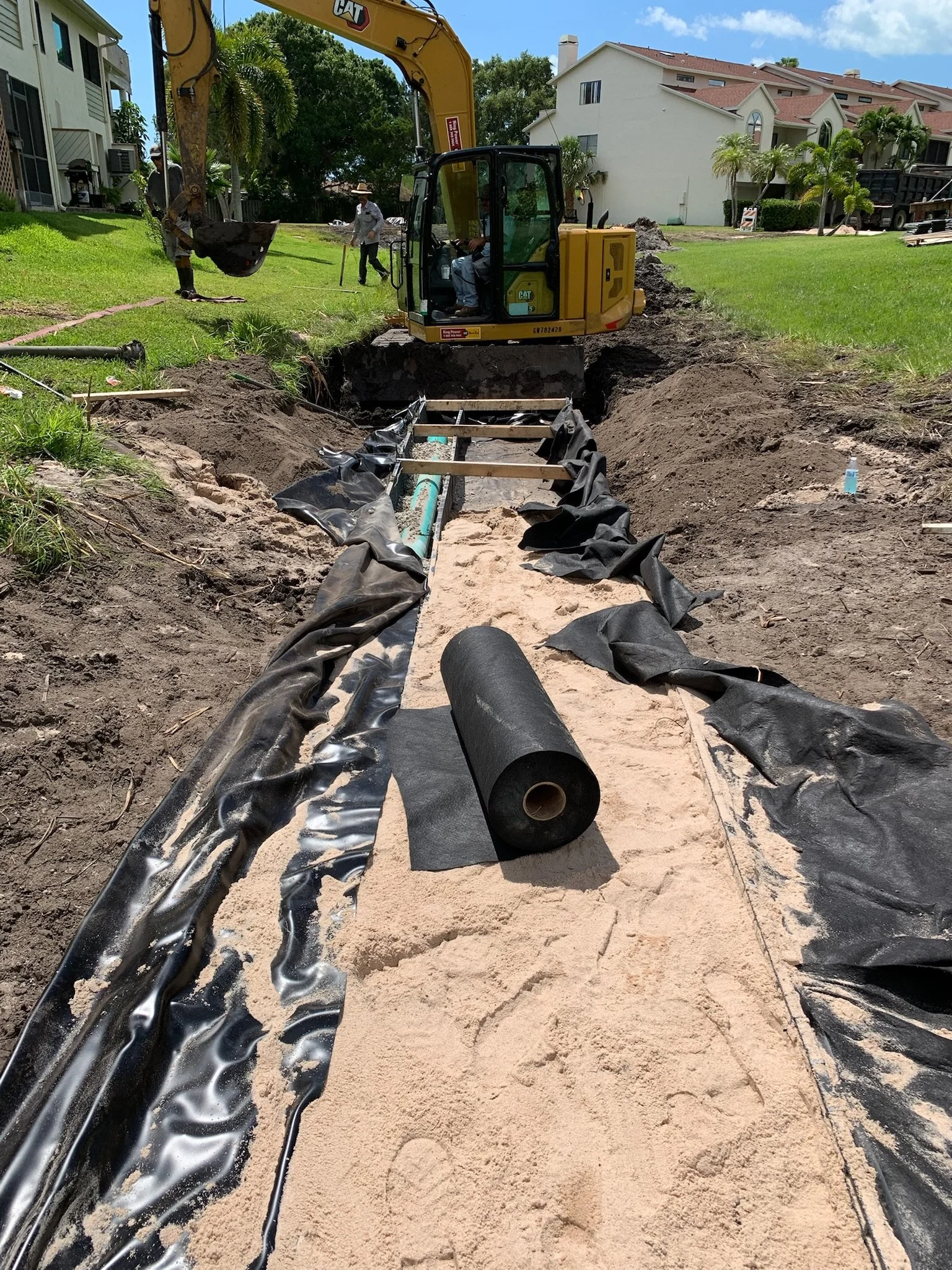 Construction workers installing underground piping with an excavator in a residential area. Specialized under drain system.