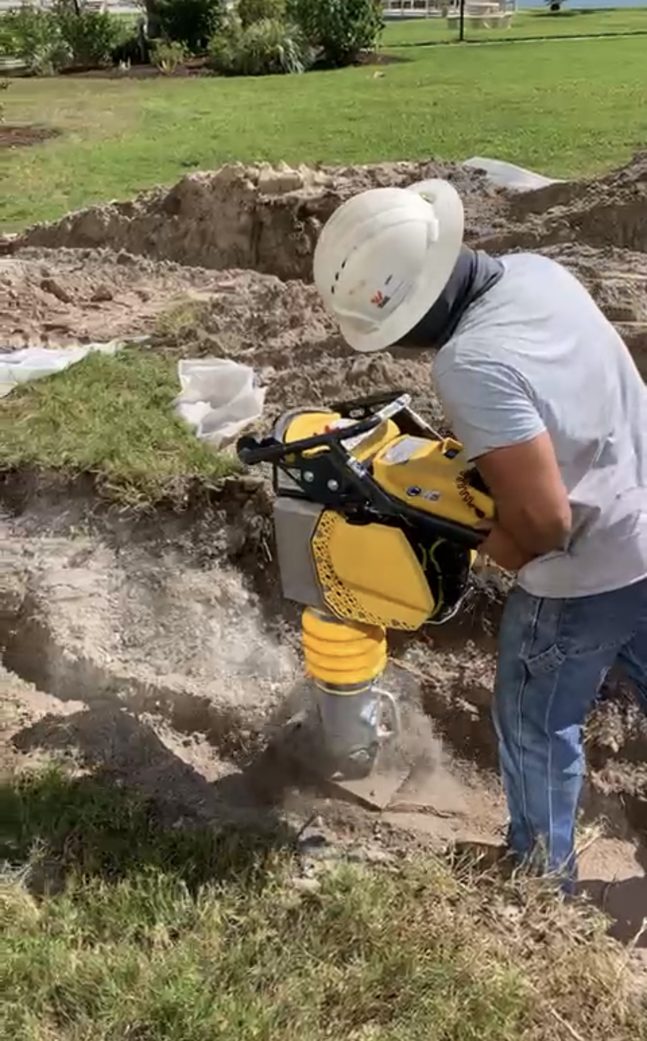 A construction worker wearing a white hard hat and white t-shirt operates a yellow compactor on the ground in a grassy area, with a dug trench nearby.