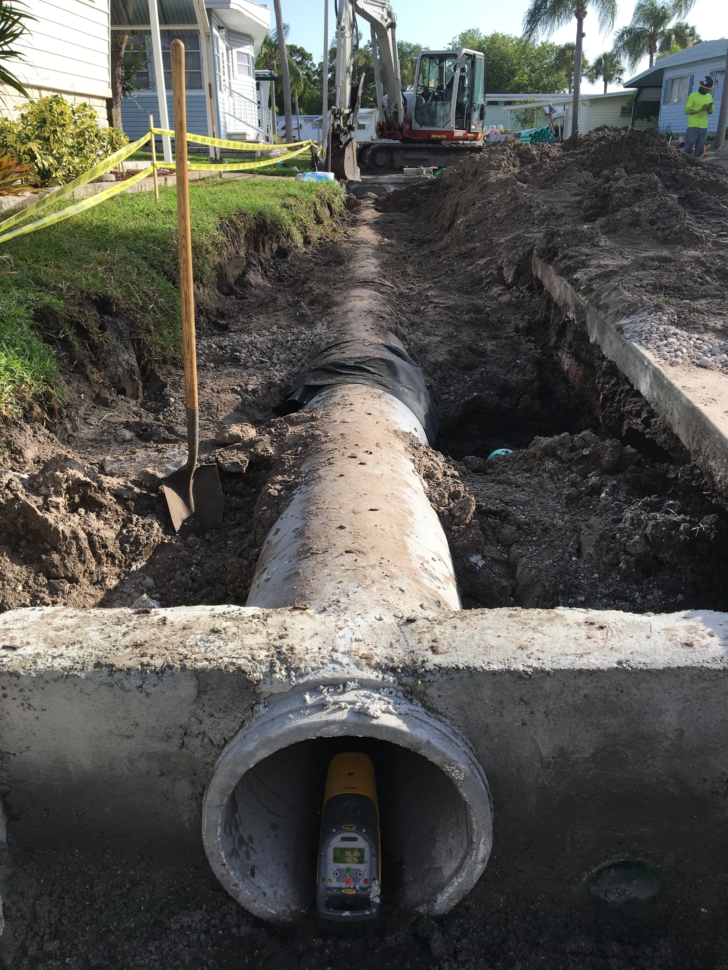 A construction site showing an excavation of a trench for RCP installation. A large pipe is in place, with a digital leveling laser inside it. There's a small excavator in the background, along with construction workers, houses, and palm trees.