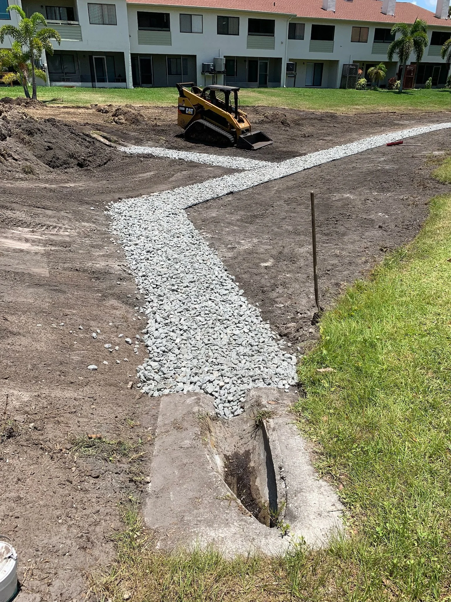 A construction site with a small Caterpillar skid-steer loader and a trench with gravel, grass, and dirt surrounding it, in front of an apartment building.