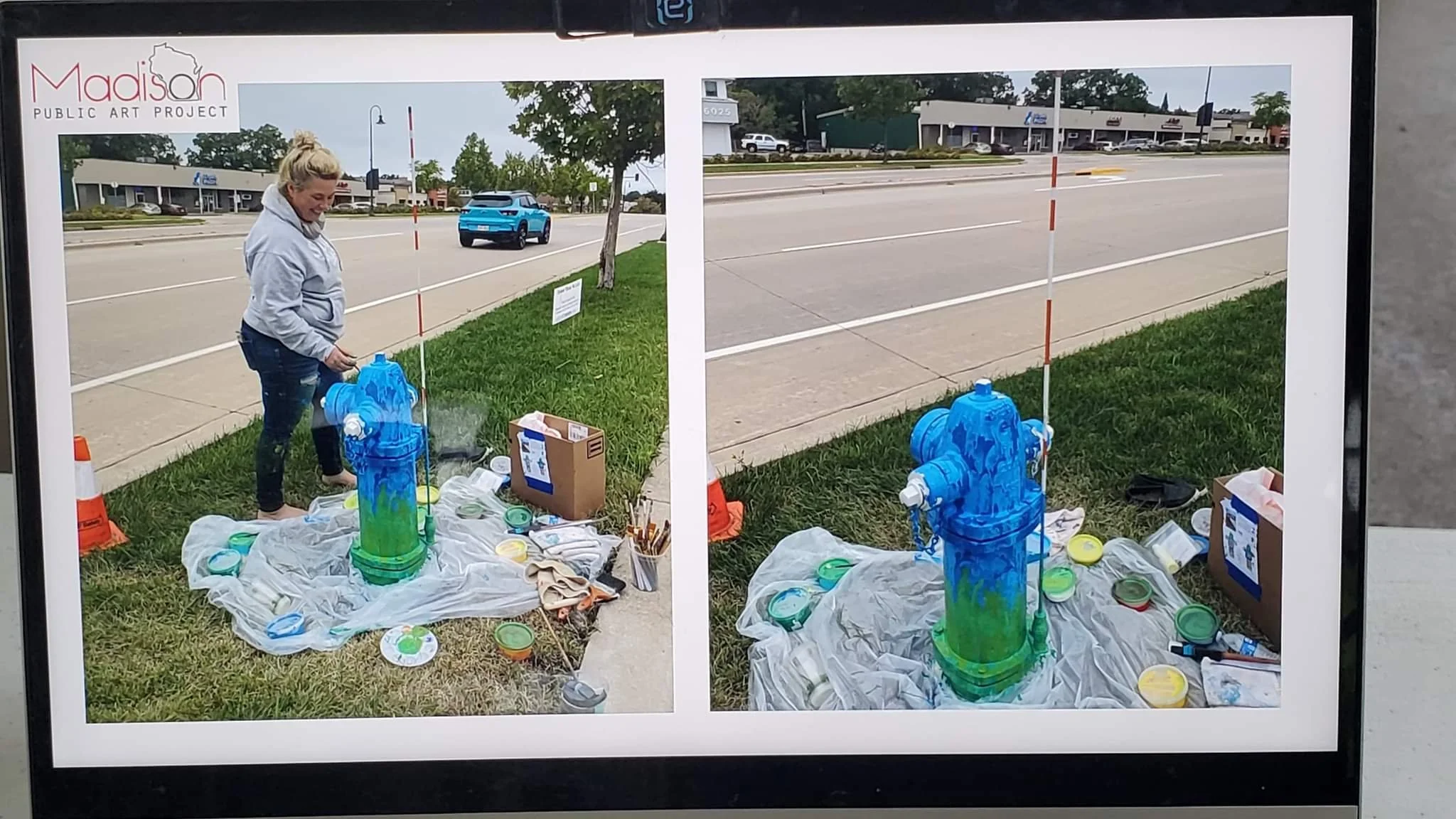 Photograph of a person painting a water bottle sculpture outdoors, with paint supplies and a box nearby, in front of a street and shopping plaza.