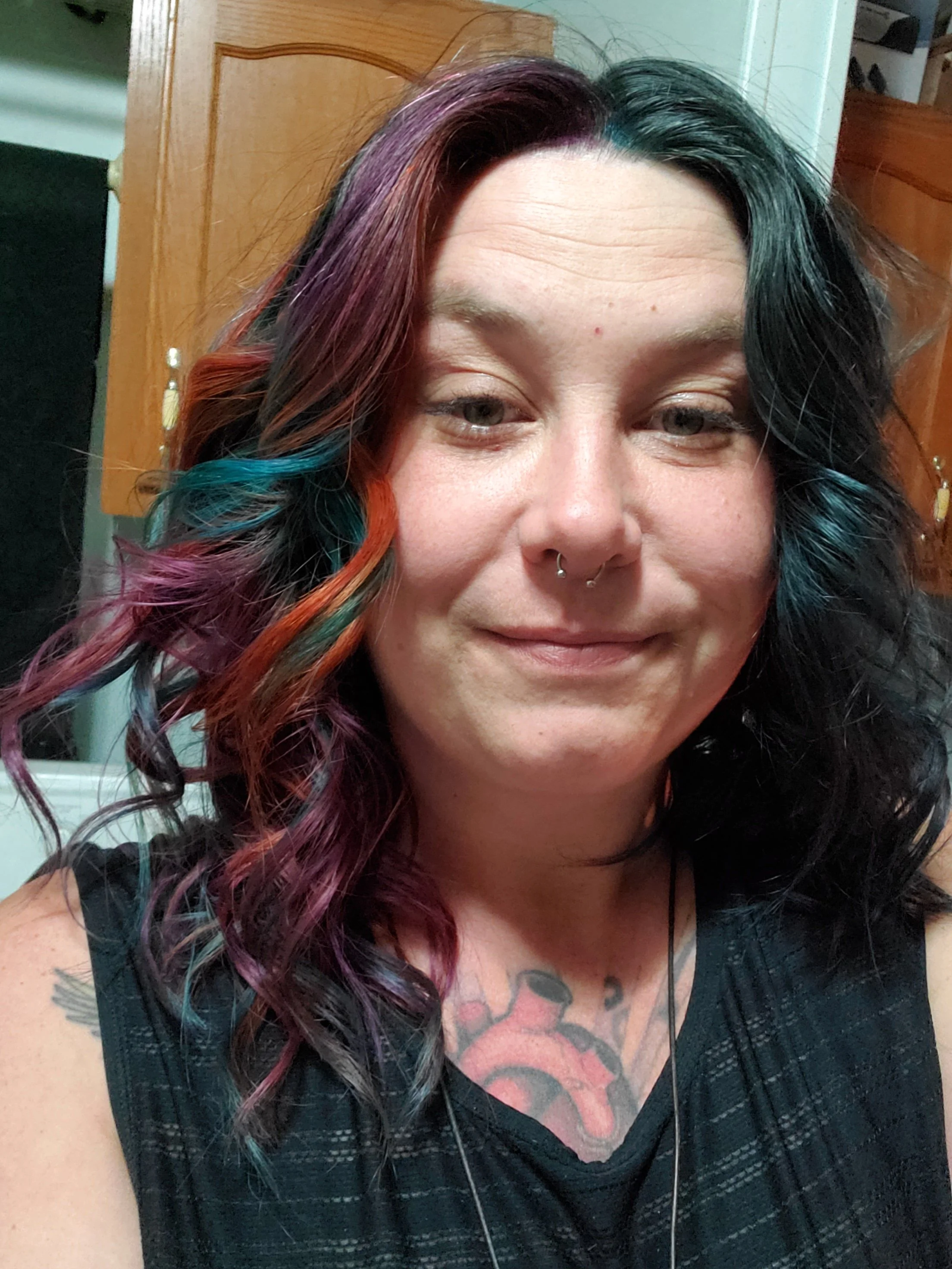A woman with long, curly hair dyed in multiple colors, including purple, pink, orange, green, and blue, smiling and wearing a black top with tattoos visible on her chest.
