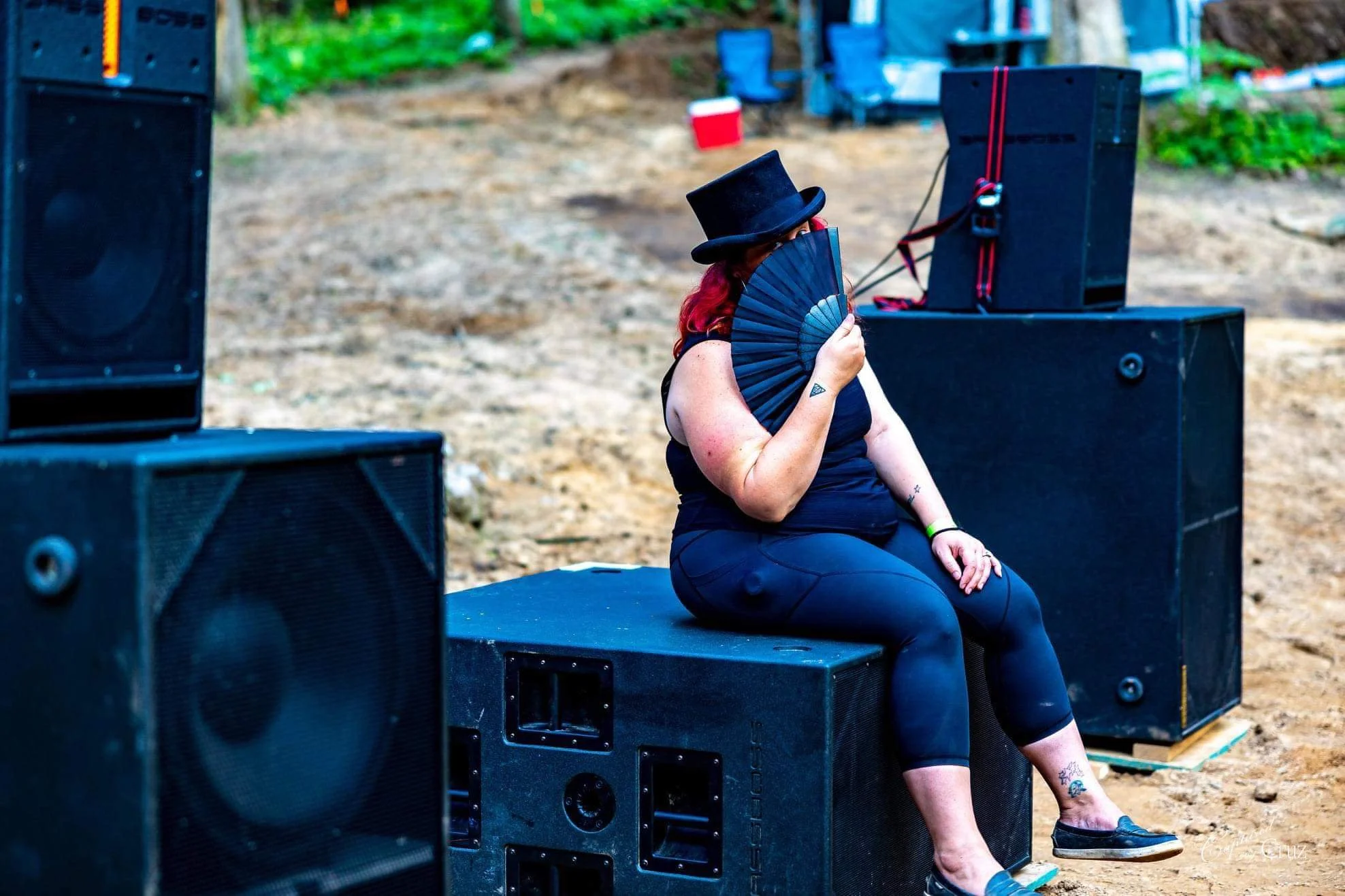A woman with red hair, tattoos, and a black top hat is sitting on a black stage or platform outdoors, holding a black fan in front of her face. She is wearing black clothing and has piercings. Large black speakers are positioned around her, indicating a music or event setting.