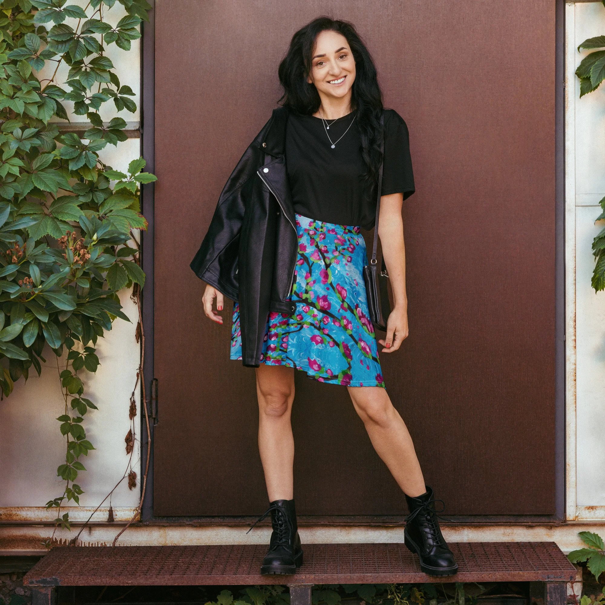 A young woman with long black hair, wearing a black t-shirt, colorful patterned skirt, black lace-up boots, and a black leather jacket draped over her shoulder, smiling while standing outdoors in front of a brown wall with green leafy plants on either side.