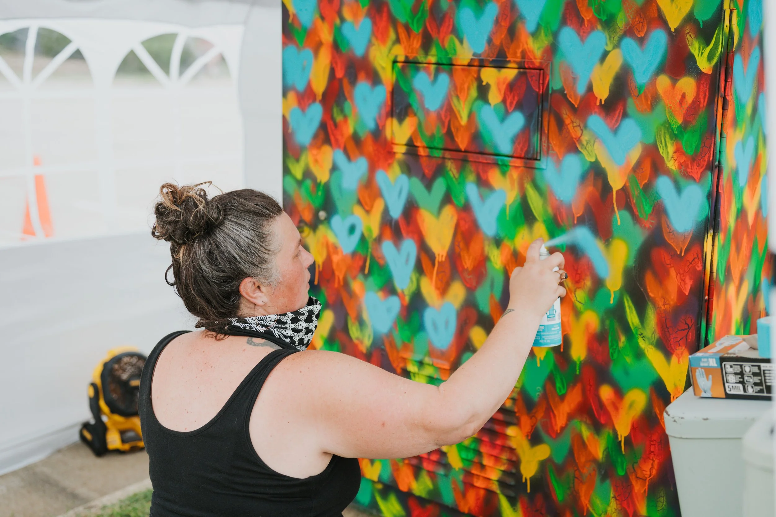 A woman with curly brown hair in a bun, wearing a black tank top and a black and white face mask, sprays paint onto a colorful wall covered with blue, red, yellow, and green hearts.