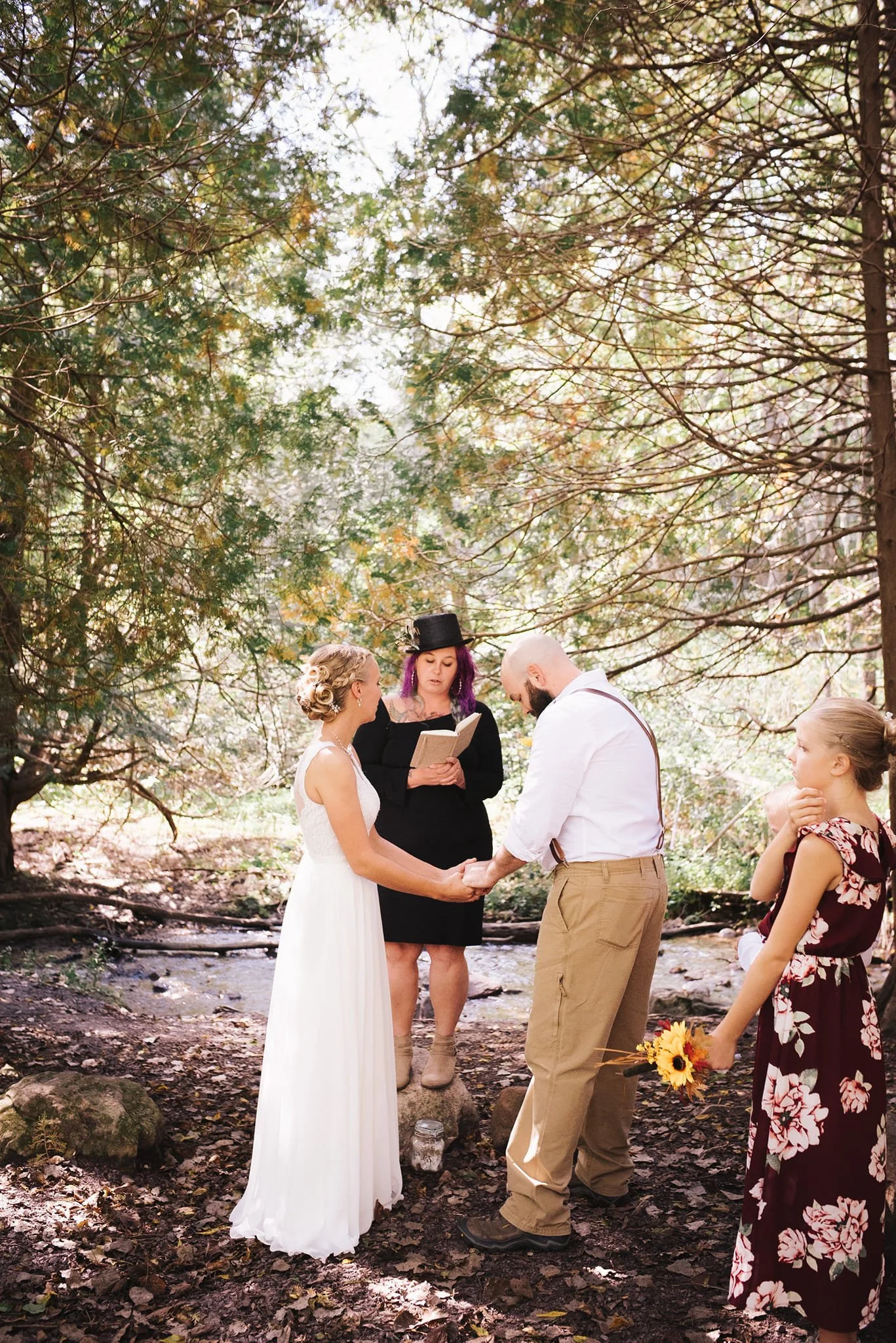 A wedding ceremony taking place outdoors in a forest, with a bride and groom holding hands and facing each other, surrounded by a small group of witnesses. The bride wears a white dress, and the groom wears tan pants and a white shirt. A woman officiates the ceremony, holding a book, with purple hair and a black hat. One woman is holding a bouquet of sunflowers, and another girl is standing nearby, dressed in a floral dress.