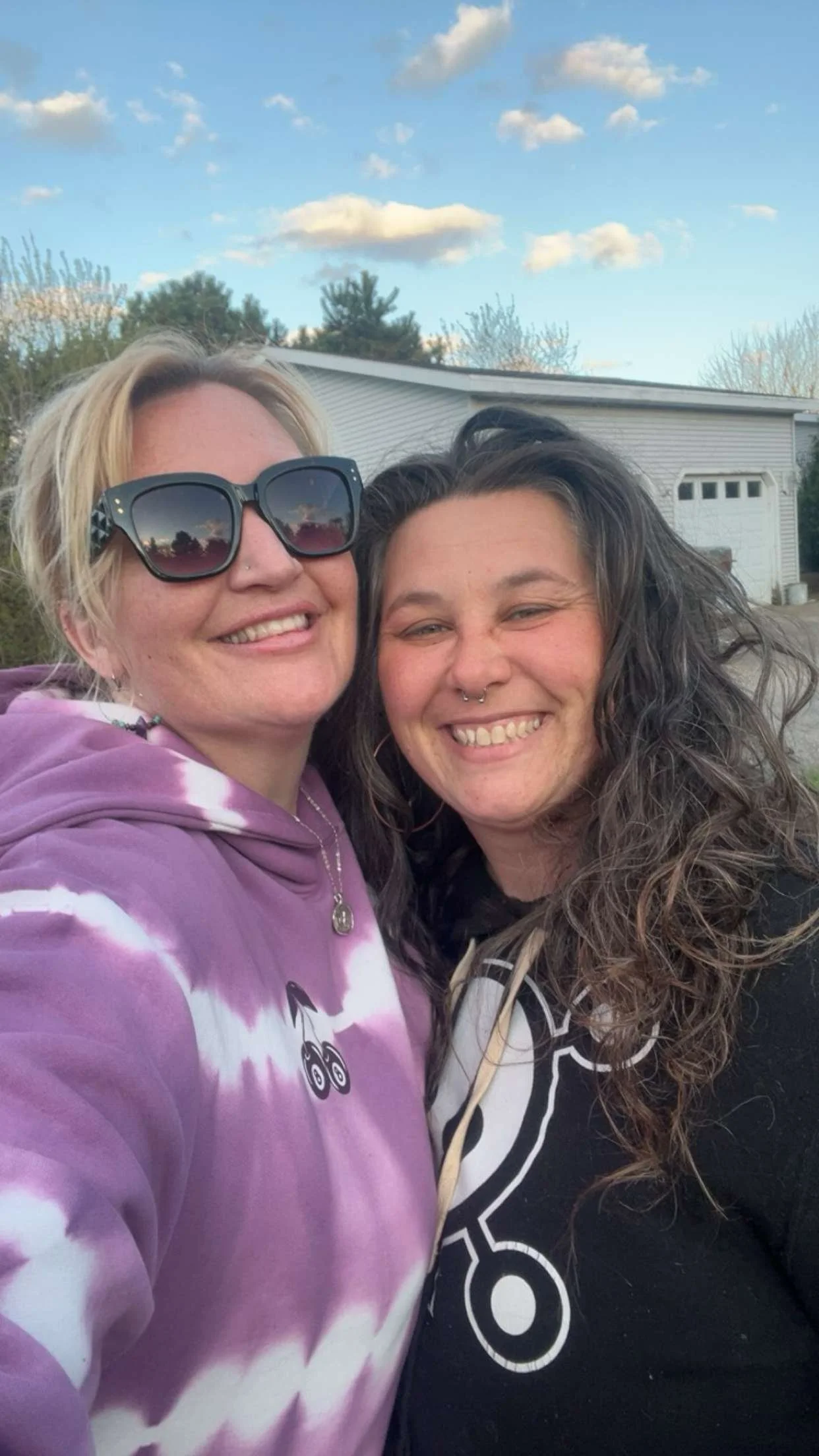 Two women- Sarah Hoekstra & Jessica Fuchs - smiling for a selfie outdoors, with a house, trees, and a partly cloudy sky in the background.