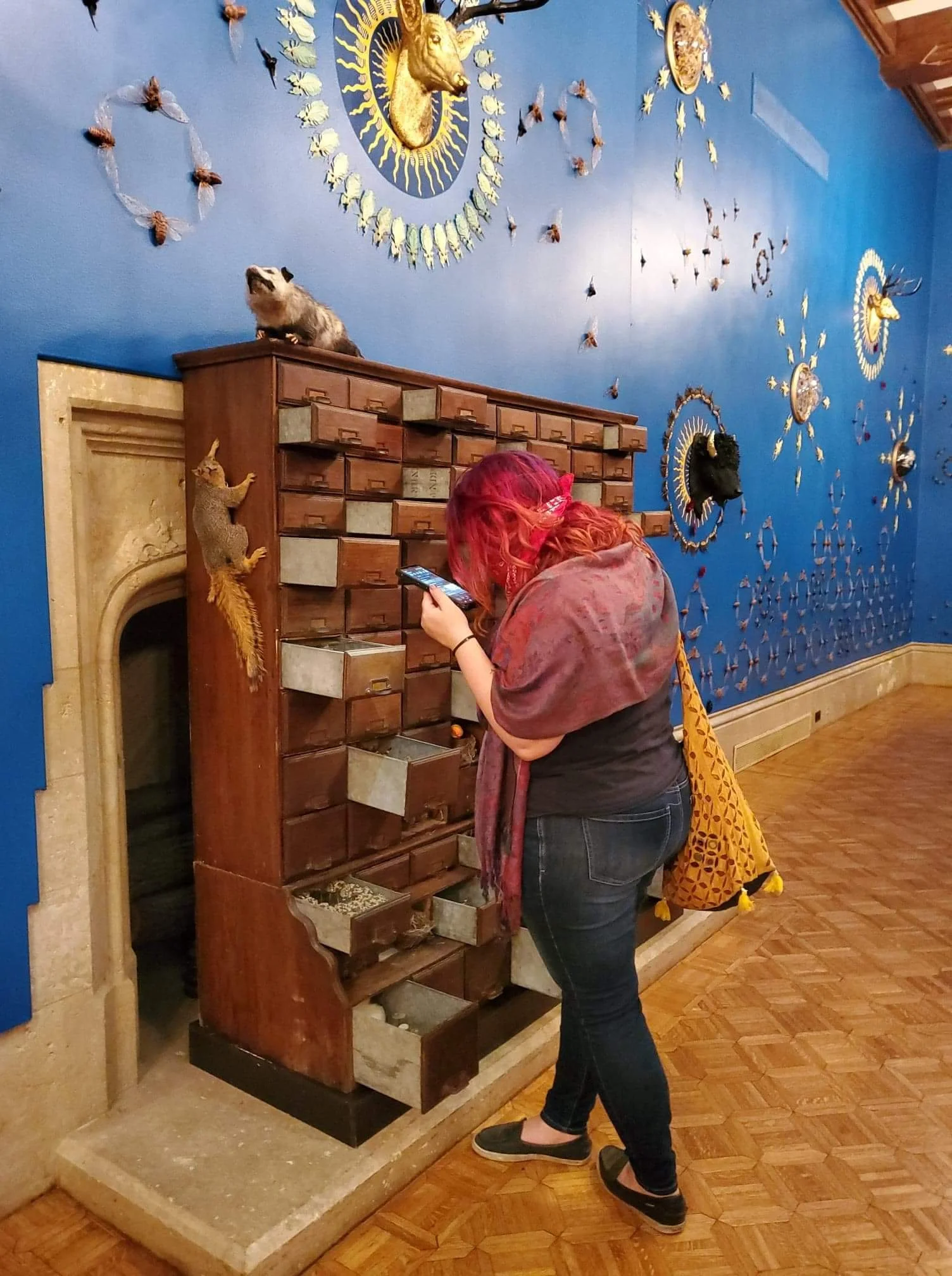A woman with red hair, wearing a scarf and jeans, looks at a vintage wooden key cabinet with multiple small drawers. She is in a room with a blue wall decorated with animal mountings and butterflies, and a person wearing a yellow patterned shirt is peeking from behind the cabinet.