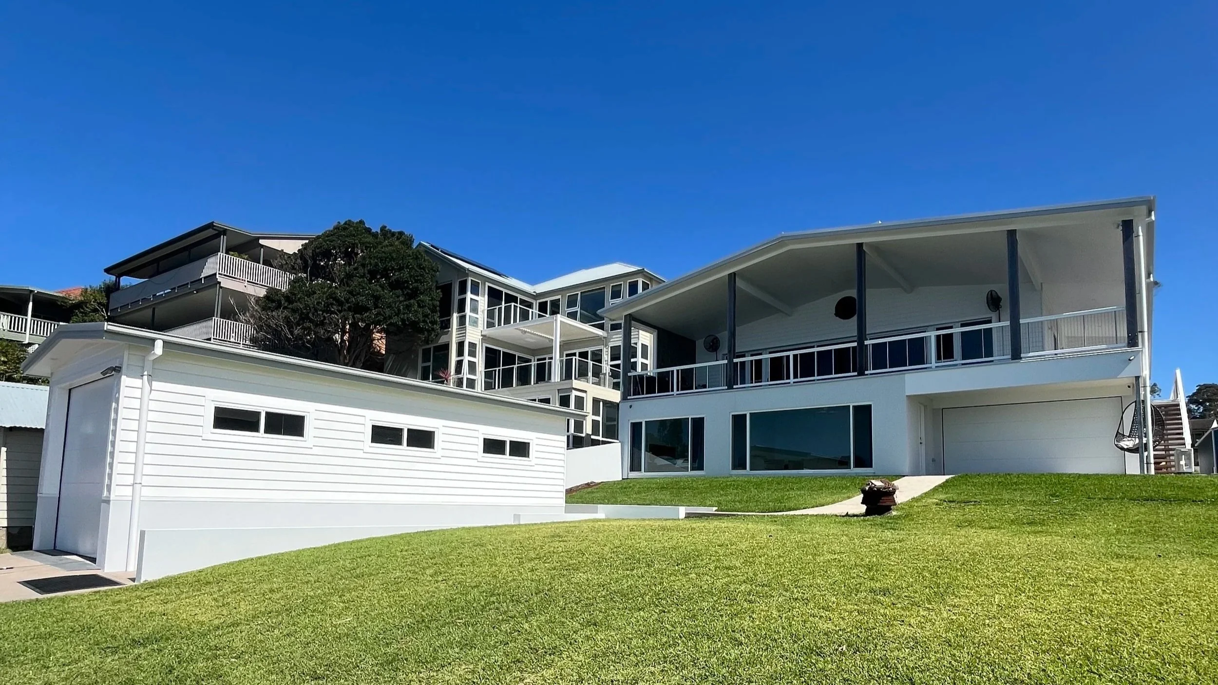 Modern white multi-story house with large windows, balconies, and a well-maintained green lawn under a clear blue sky.