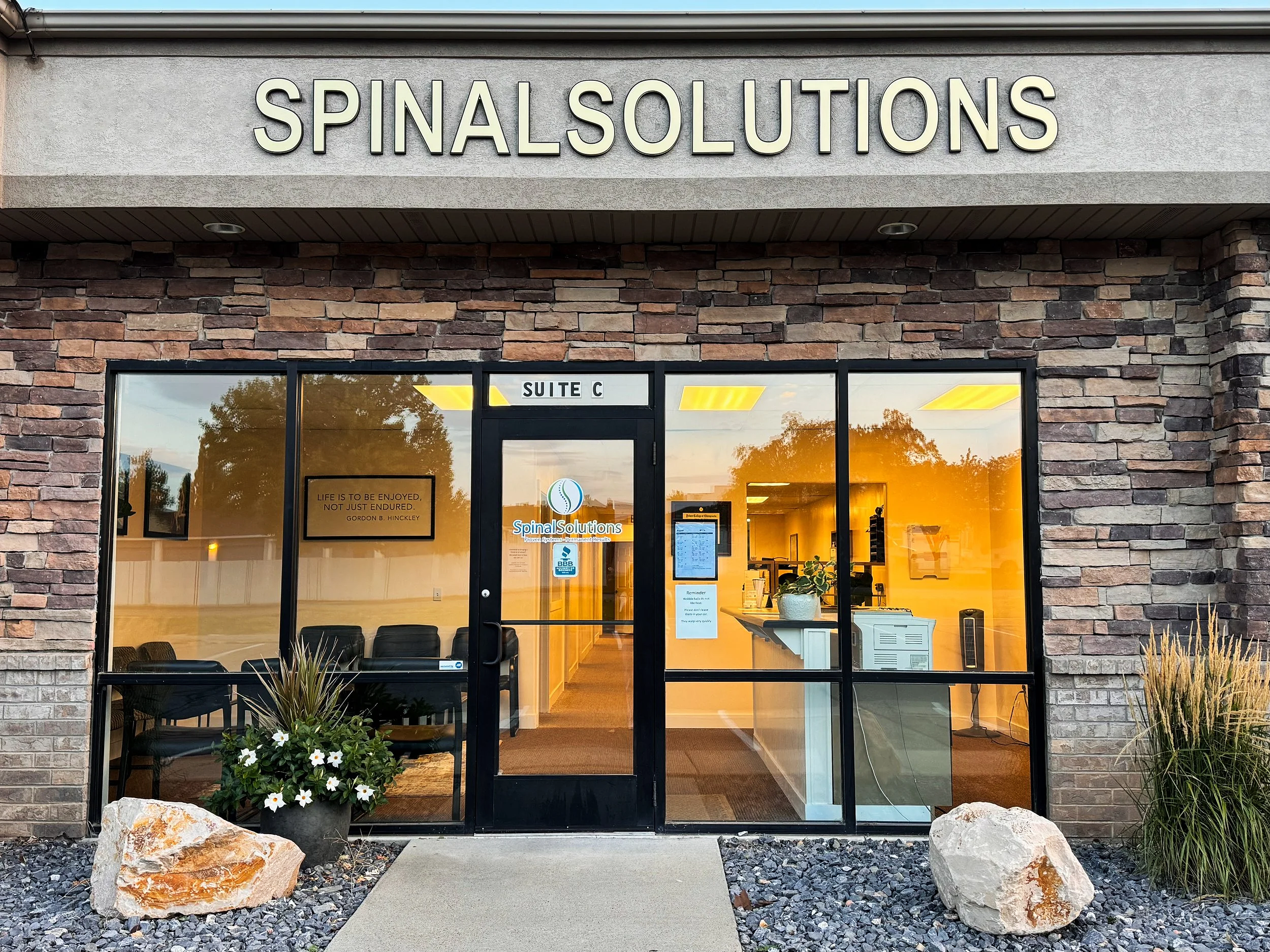 Exterior view of a building with a sign that reads 'Spinal Solutions'. The entrance features glass doors and windows, with potted plants and rocks in front. Inside, chairs and a reception area are visible.