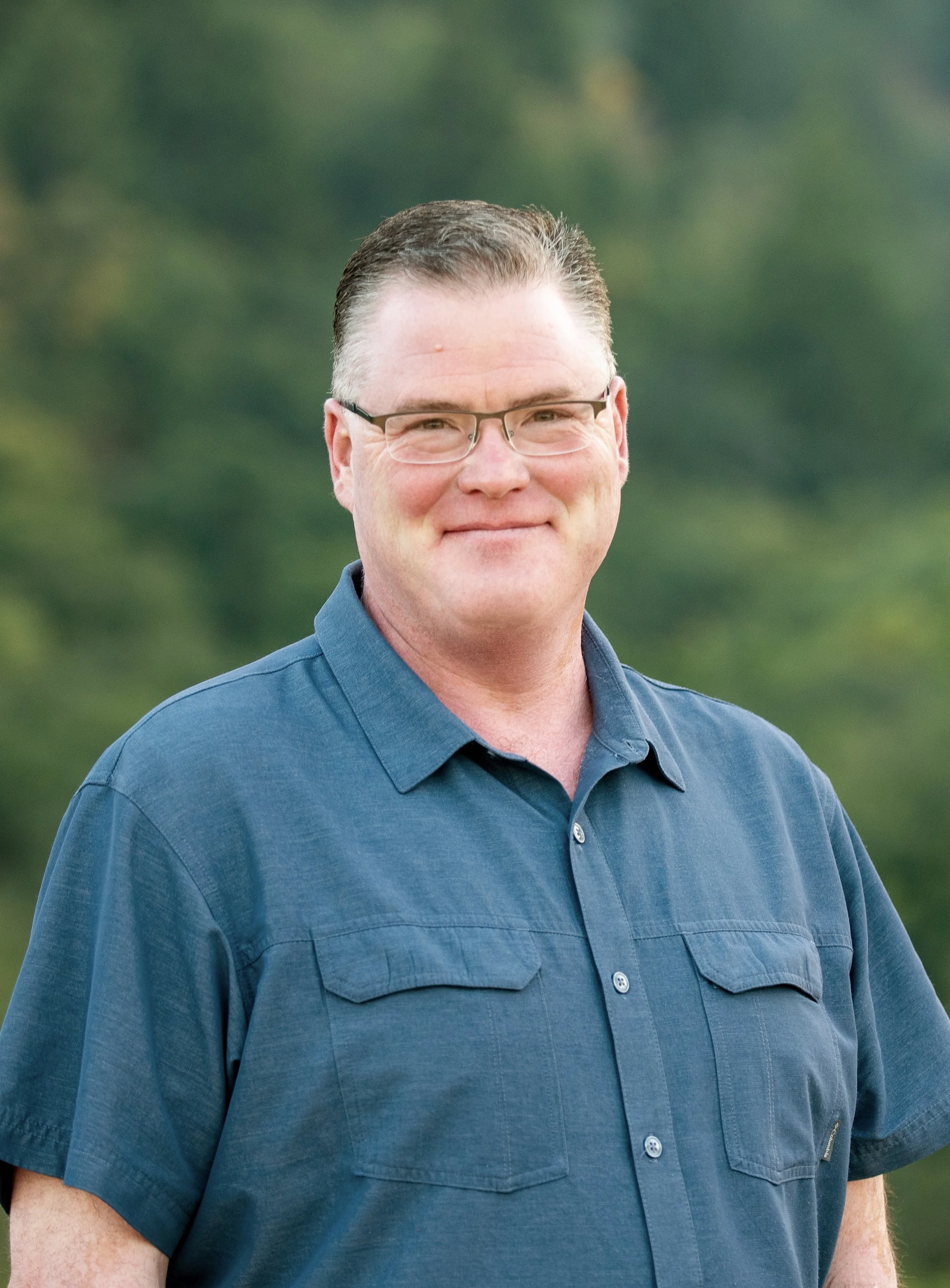A middle-aged man with glasses, short dark hair, and a light complexion wearing a blue button-up shirt, outdoors with greenery in the background.