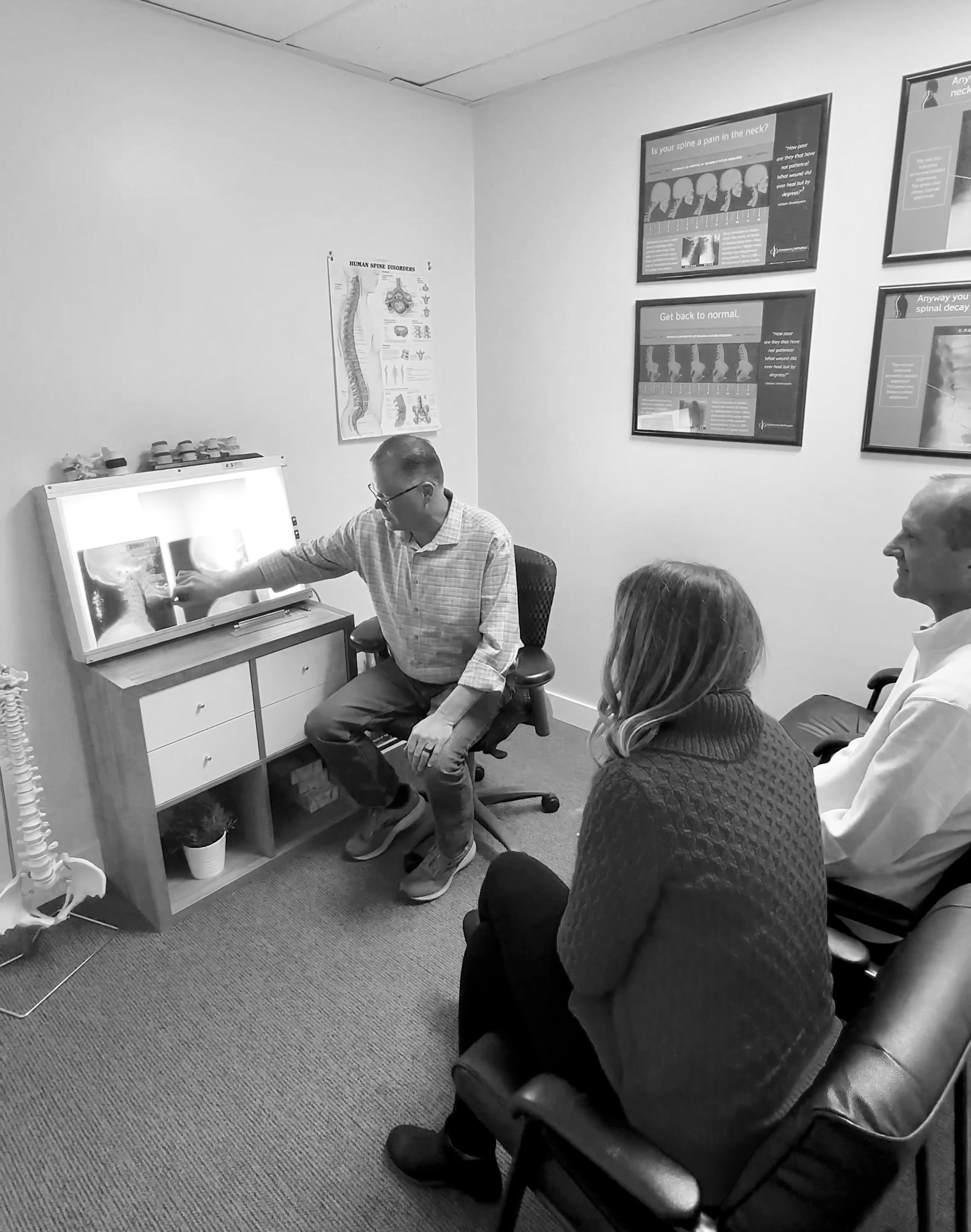 A group of people sitting in a room, watching a presentation or demonstration about a medical or anatomical topic related to the human spine, with a man in a checkered shirt sitting in front of a mirror that displays some of his back.