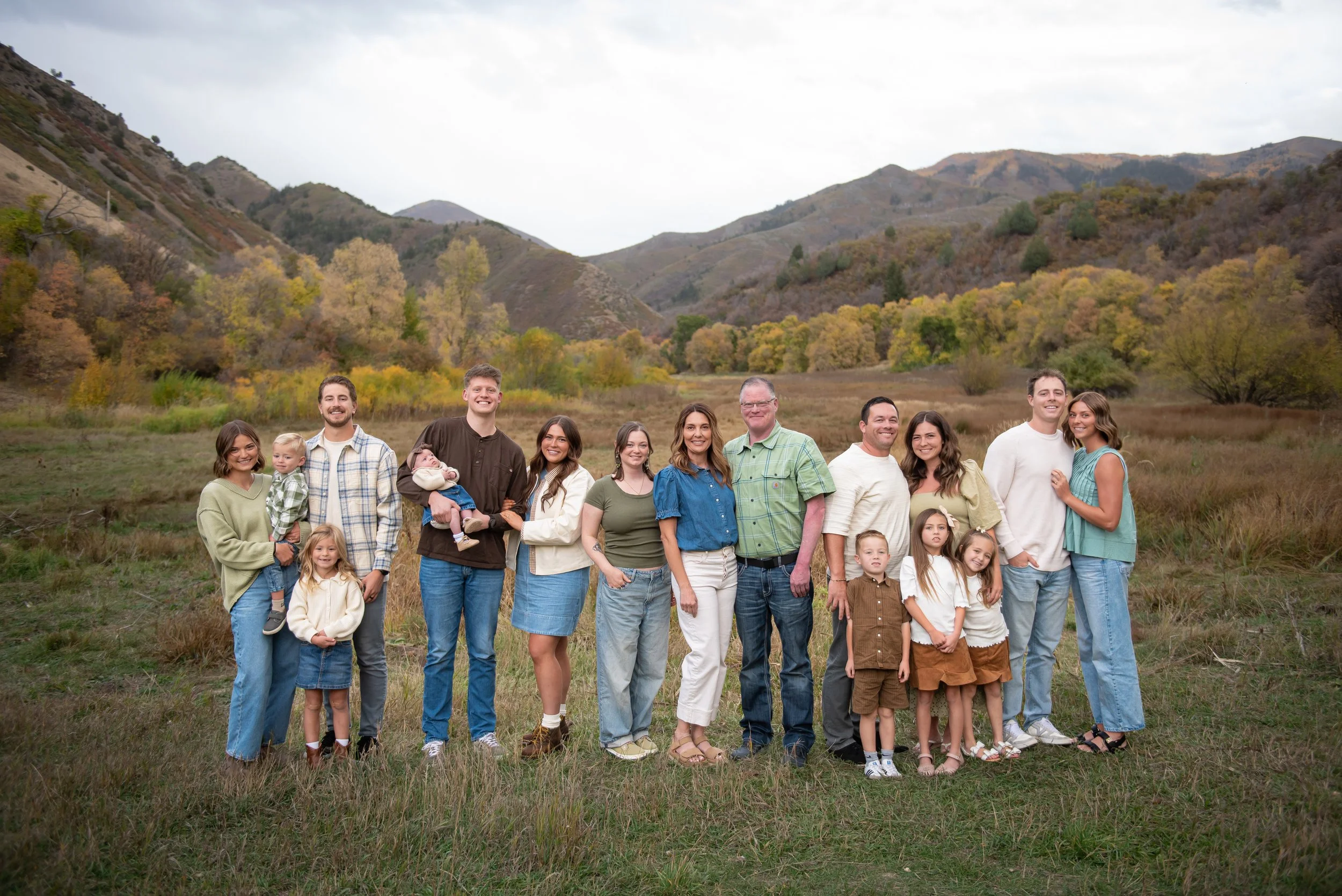 A large group of people, including children and adults, standing outdoors in a grassy field with mountains and trees in the background. The sky is overcast.