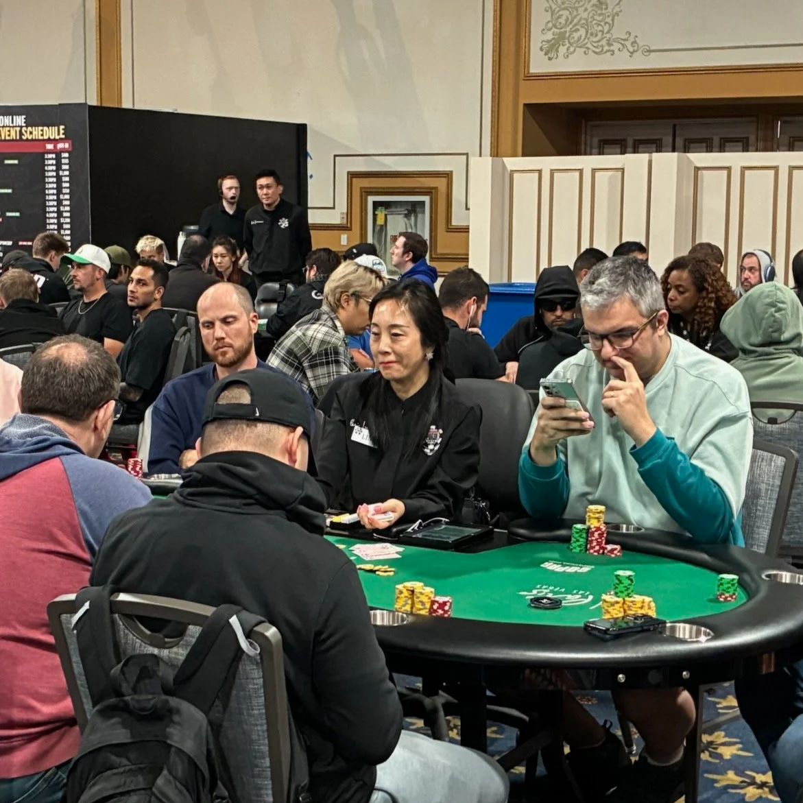 People playing poker at a casino table with chips, surrounded by other players, in a crowded room with a welcome sign and staff in the background.