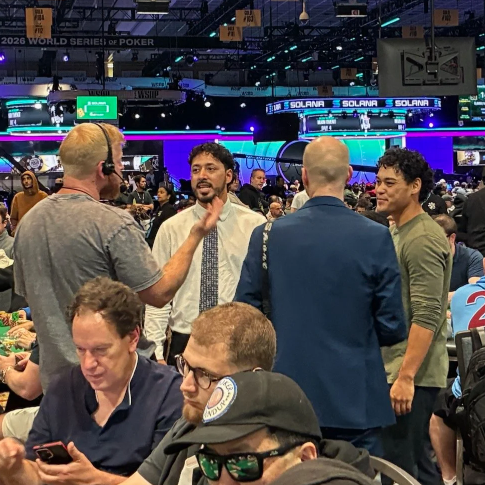 A crowded scene at a poker tournament with people engaged in conversations, some seated at poker tables and some standing. Bright neon lights and screens are visible in the background, indicating a lively gaming environment.
