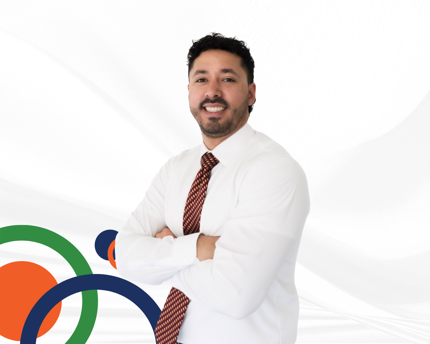 A man with dark hair and a beard wearing a white shirt and patterned tie, standing with arms crossed against a white background with colorful circular design elements.
