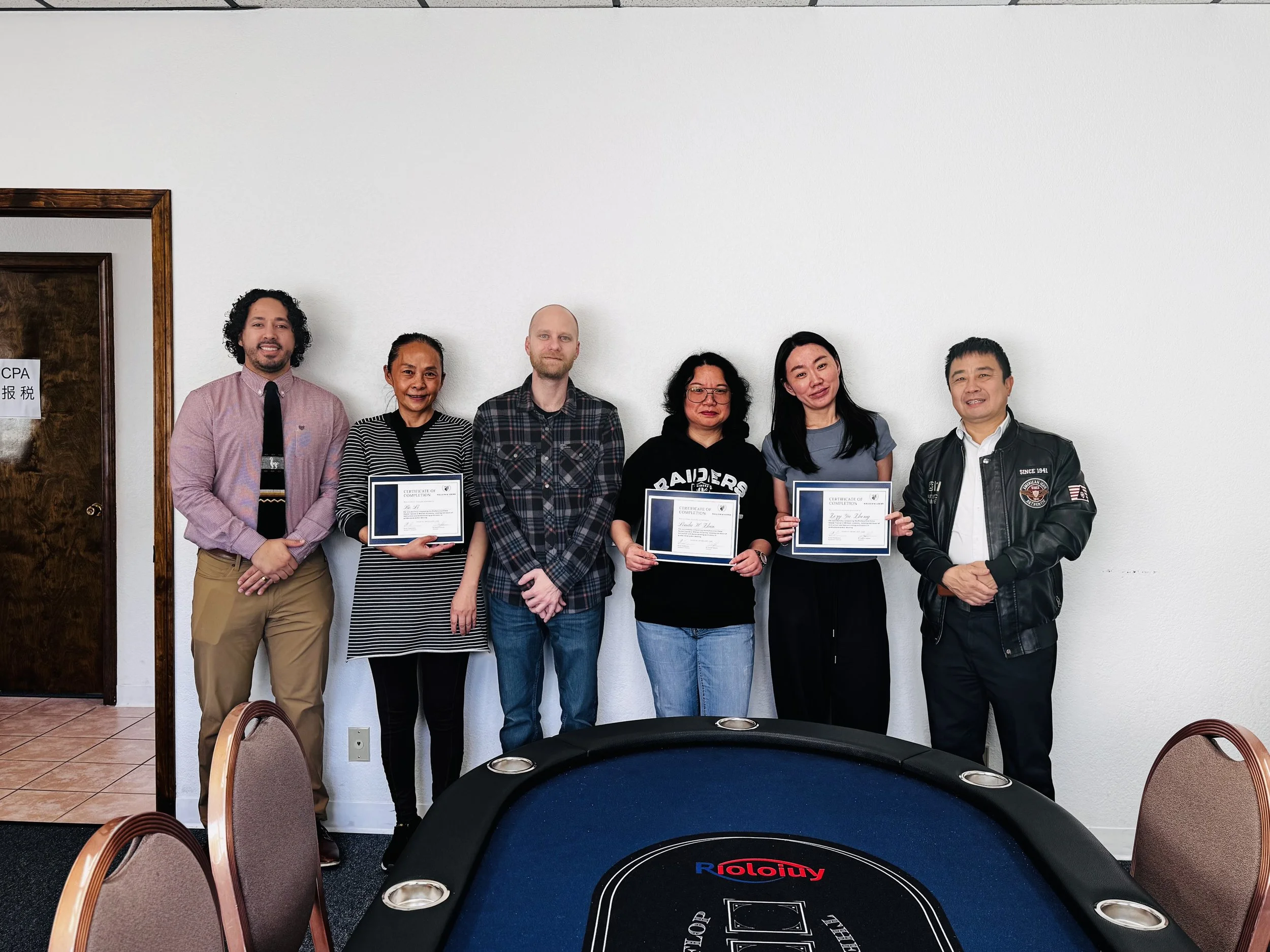 Group of six diverse people standing indoors against a white wall, some holding certificates, in a casual or semi-formal setting.