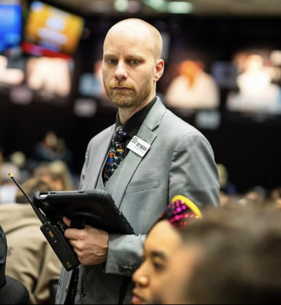 A man with a beard and shaved head wearing a gray suit and floral tie, holding a tablet device, standing in a busy environment with multiple screens in the background.
