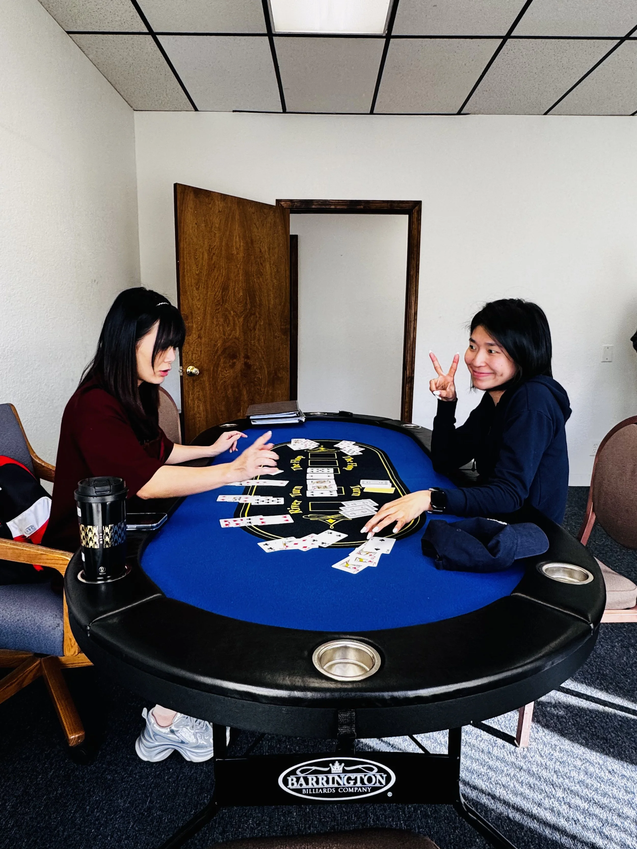 Two women playing cards at a round billiards table in a room with beige walls and a wooden door. One woman is making a peace sign and smiling, while the other is focused on her cards.