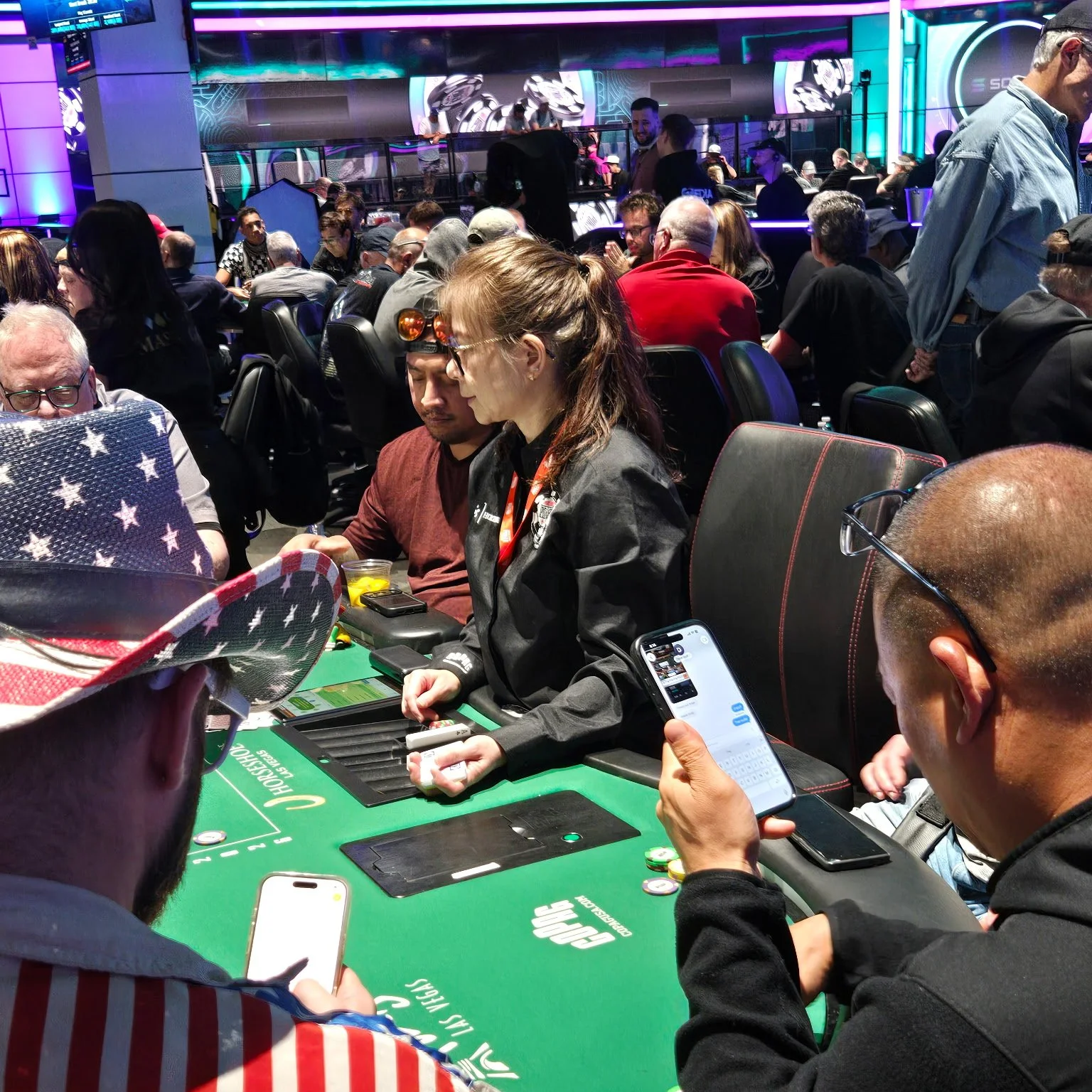 People playing poker at a casino table surrounded by a crowd in a lively gaming room with colorful lights and digital displays.