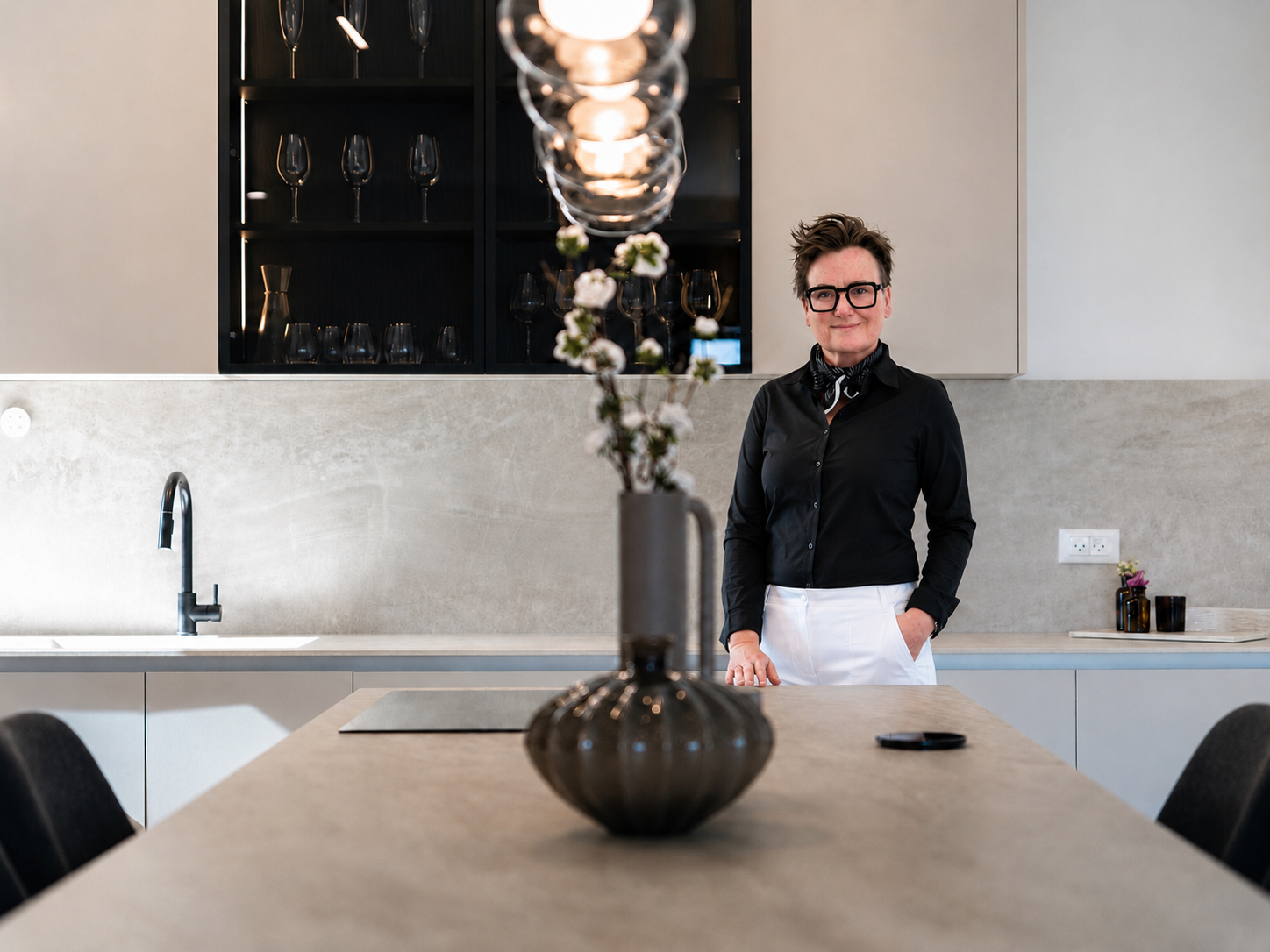 A woman with short hair, glasses, and a black shirt stands in a modern kitchen. She has her hands in her white apron pockets and is smiling slightly. The kitchen features a light-colored countertop, a sink with a black faucet, and upper shelves with glasses. A black vase with white flowers sits on the table in the foreground, and a black smartphone is on the table. The overall design is minimalistic with neutral tones.