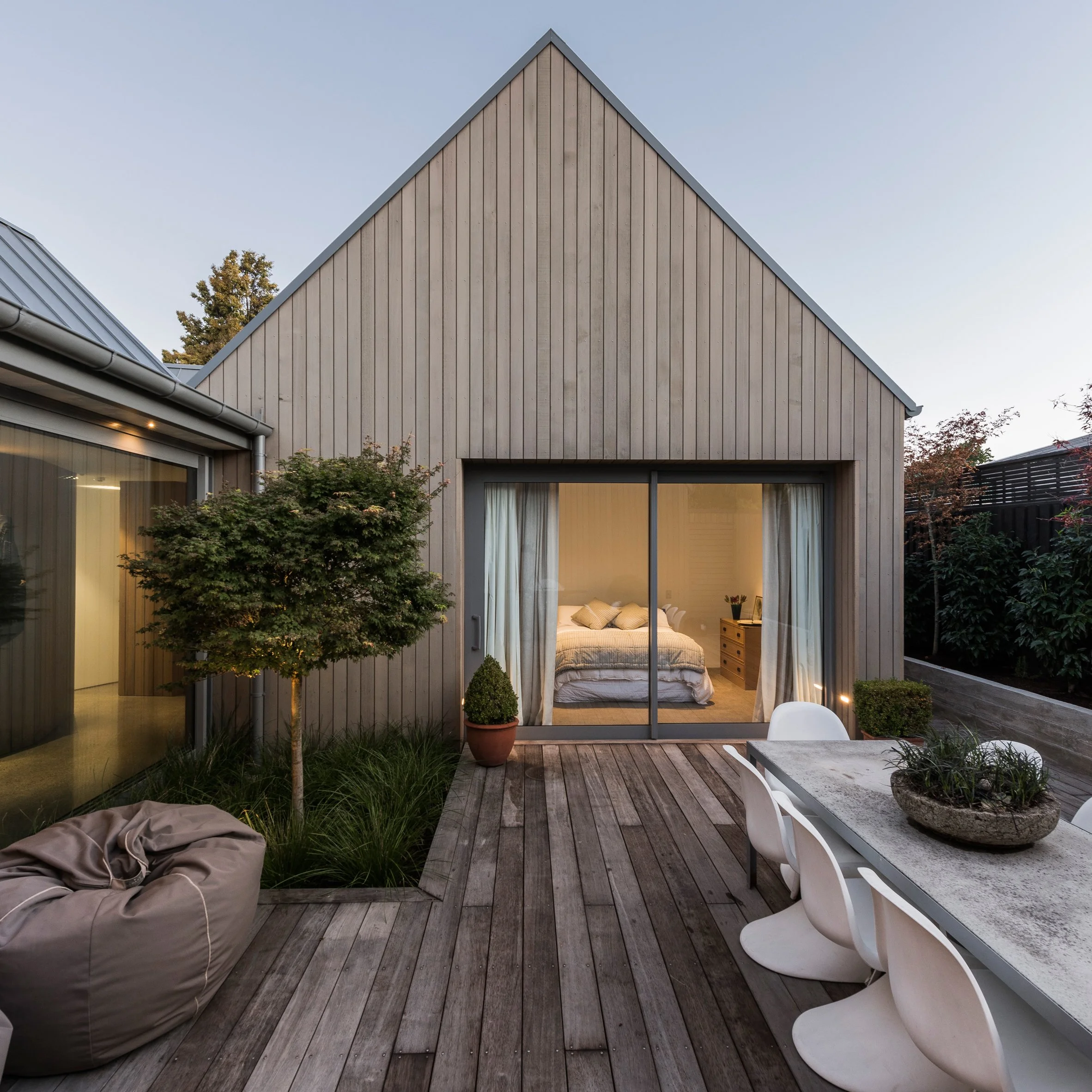 A modern house with a sharply peaked wooden facade, large windows, and a balcony, set in a landscaped backyard with trees and outdoor furniture during evening.