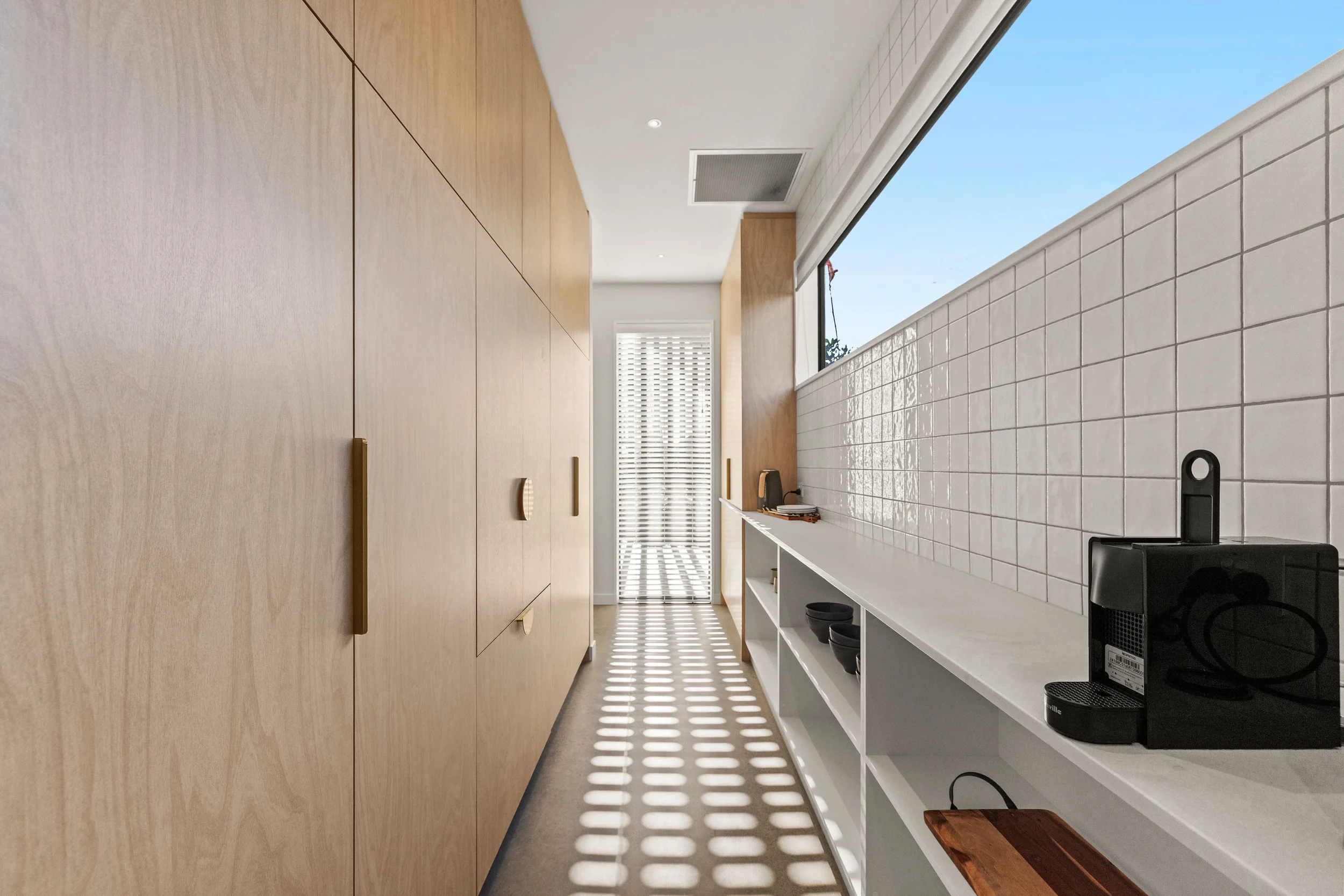 Modern kitchen with wooden cabinets on the left, white countertop on the right with black kitchen appliances, and a large window with blinds allowing sunlight to create pattern shadows on the floor.