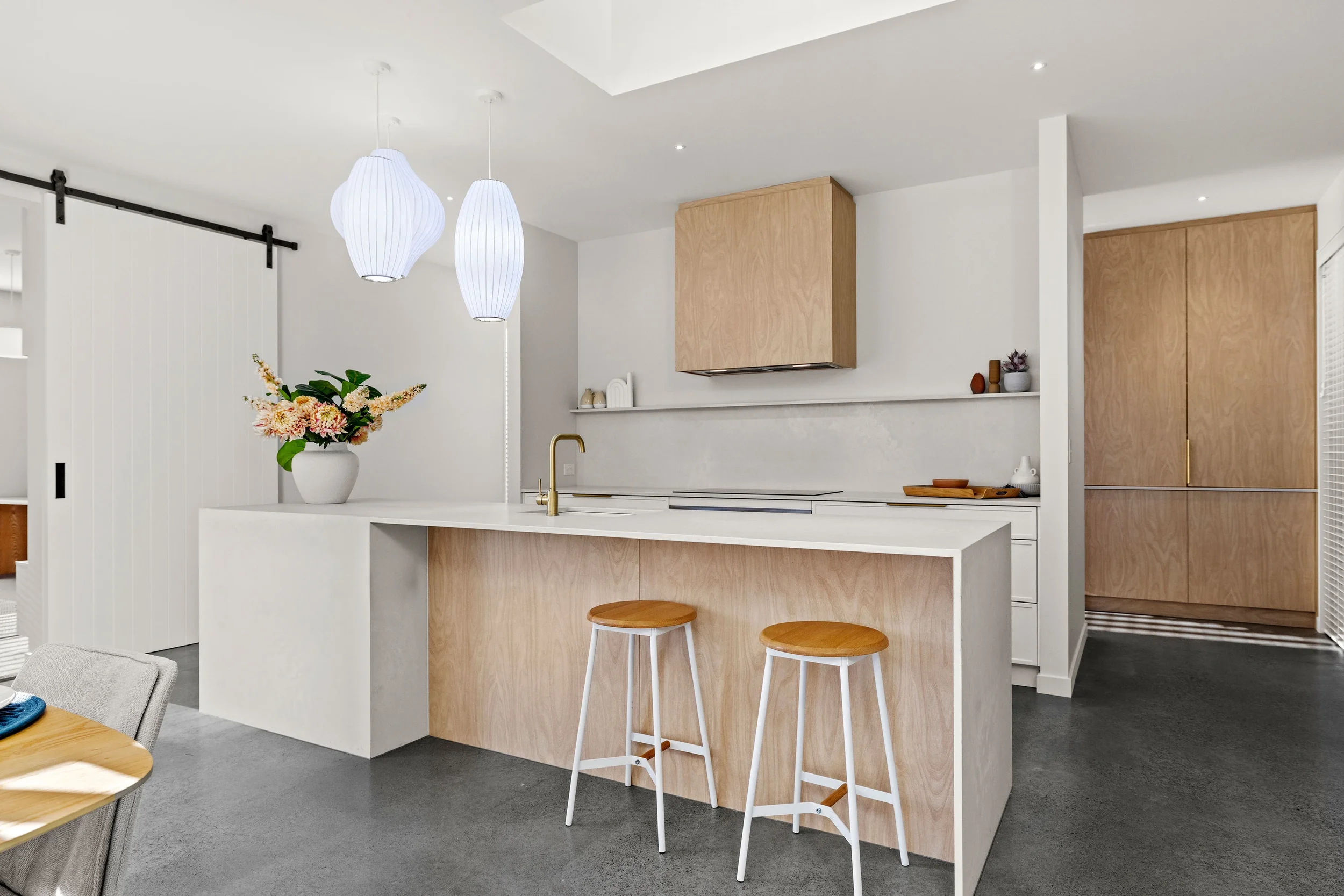 Modern kitchen with white countertops, light wood cabinets, a vase of pink and peach flowers, two white bar stools, and pendant lights with paper shades.