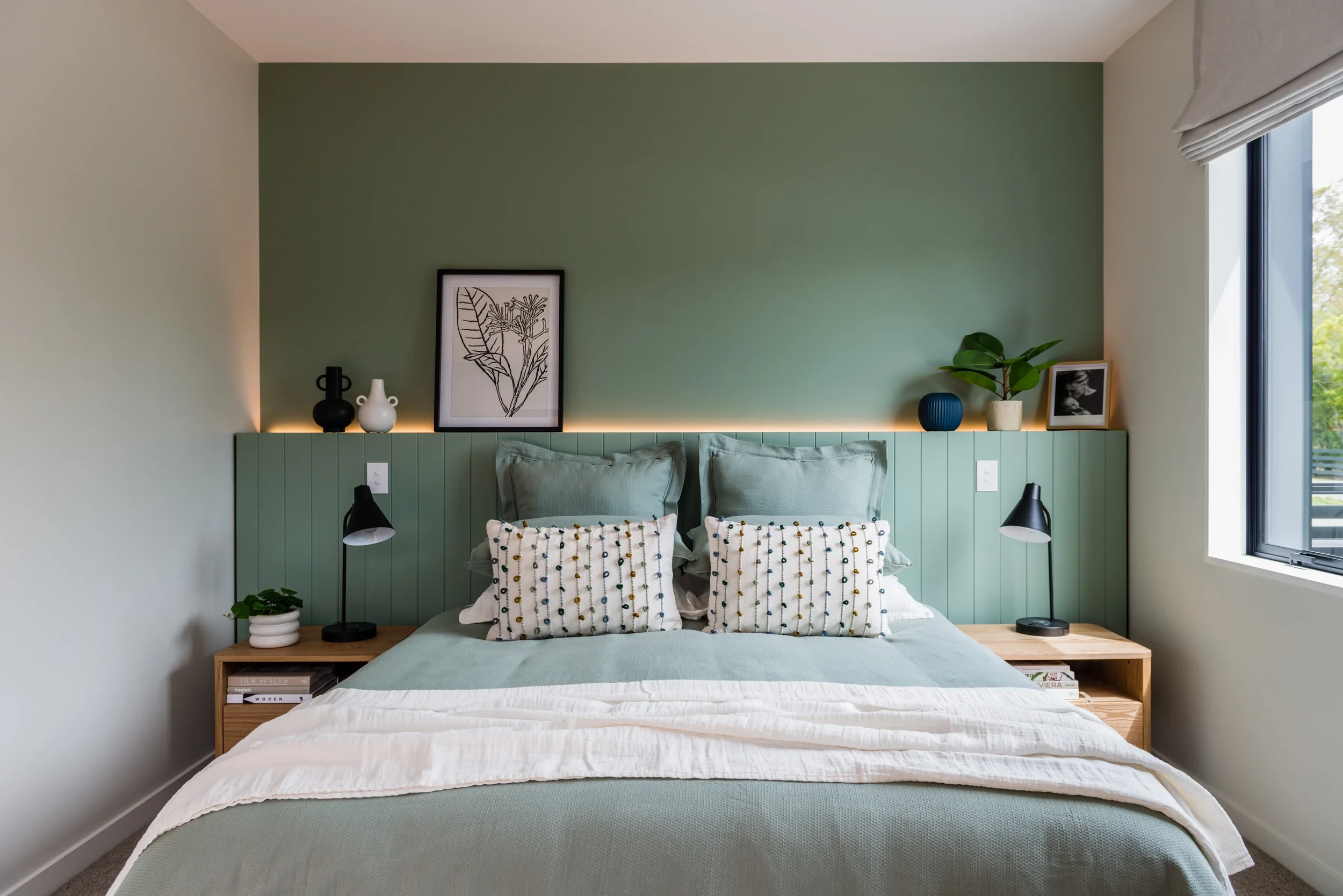 A neatly made bed with a light green bedspread and patterned pillows, against a green accent wall with black and white framed artwork, and decorative vases on a headboard and side tables, near a window with natural light.