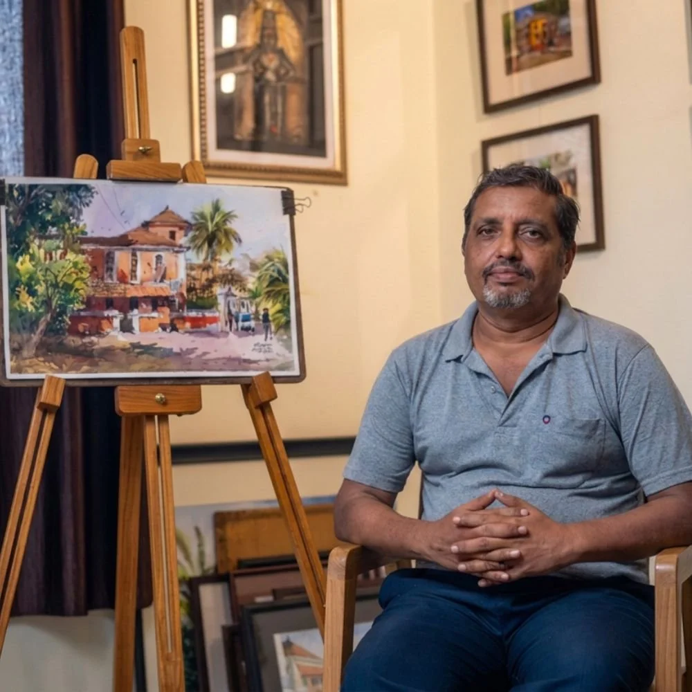 A man sitting on a wooden chair with a light blue polo shirt, in an art gallery or studio, next to an easel with a watercolor painting of a building, trees, and people, with framed artwork on the walls.