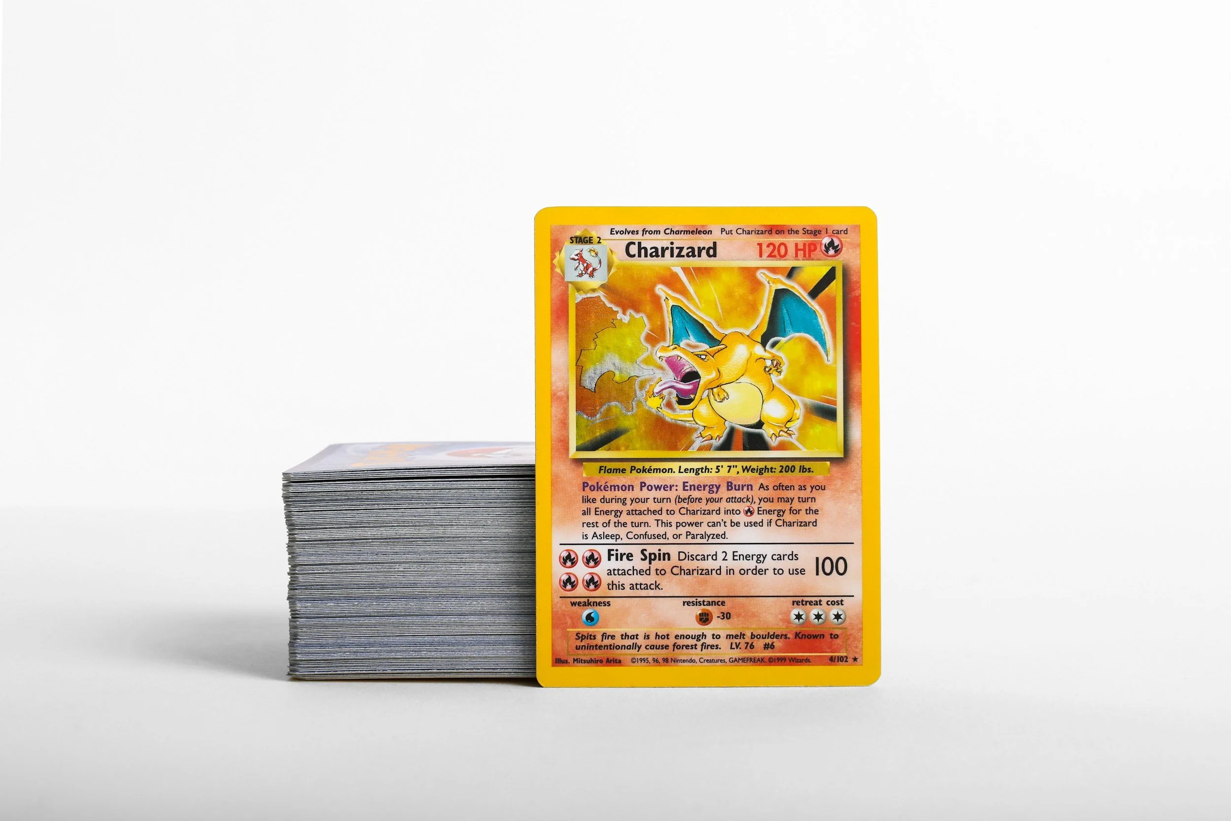 A collection of Pokémon trading cards stacked behind a single Charizard trading card in the foreground. The Charizard card features an illustration of Charizard, a fiery orange dragon, with a background of flames.