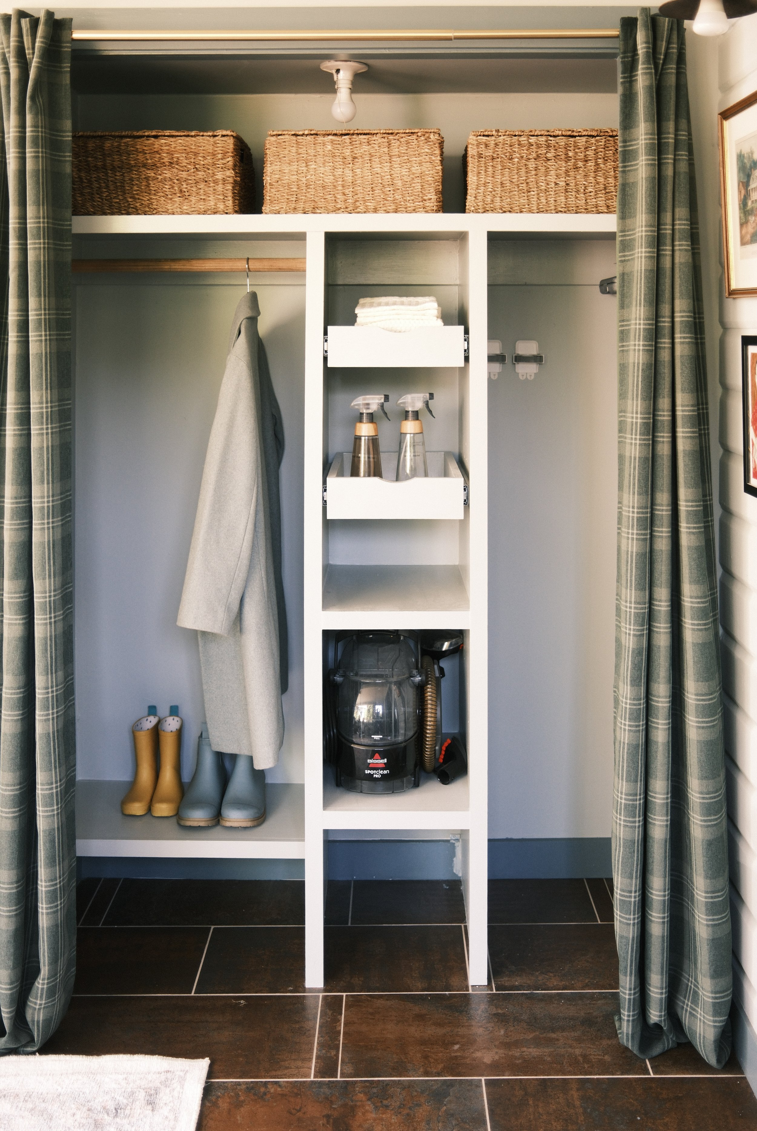 A closet with green plaid curtains pulled back, revealing a white shelving unit with baskets on top, a hanging gray coat, and shoe storage with two pairs of rain boots. There is a vacuum cleaner at the bottom of the shelving unit and spray bottles on one of the shelves.
