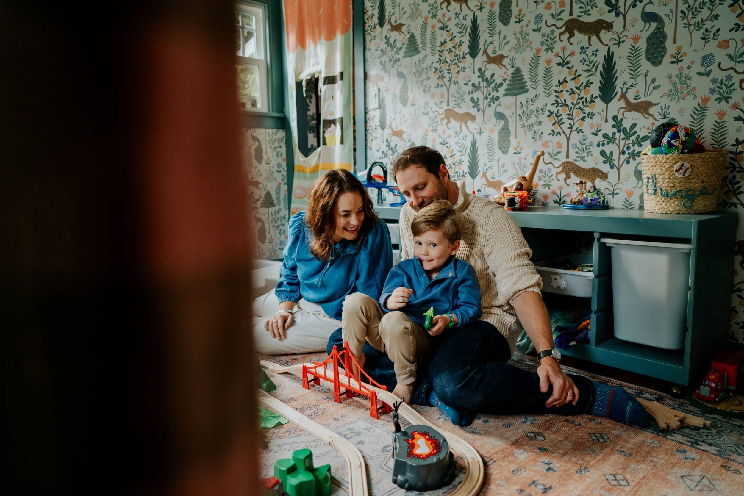 A family of three, a woman, a man, and a young boy, sitting on the floor in a child's bedroom playing with a toy train set. The room has colorful, jungle-themed wallpaper and various toys and decorations.