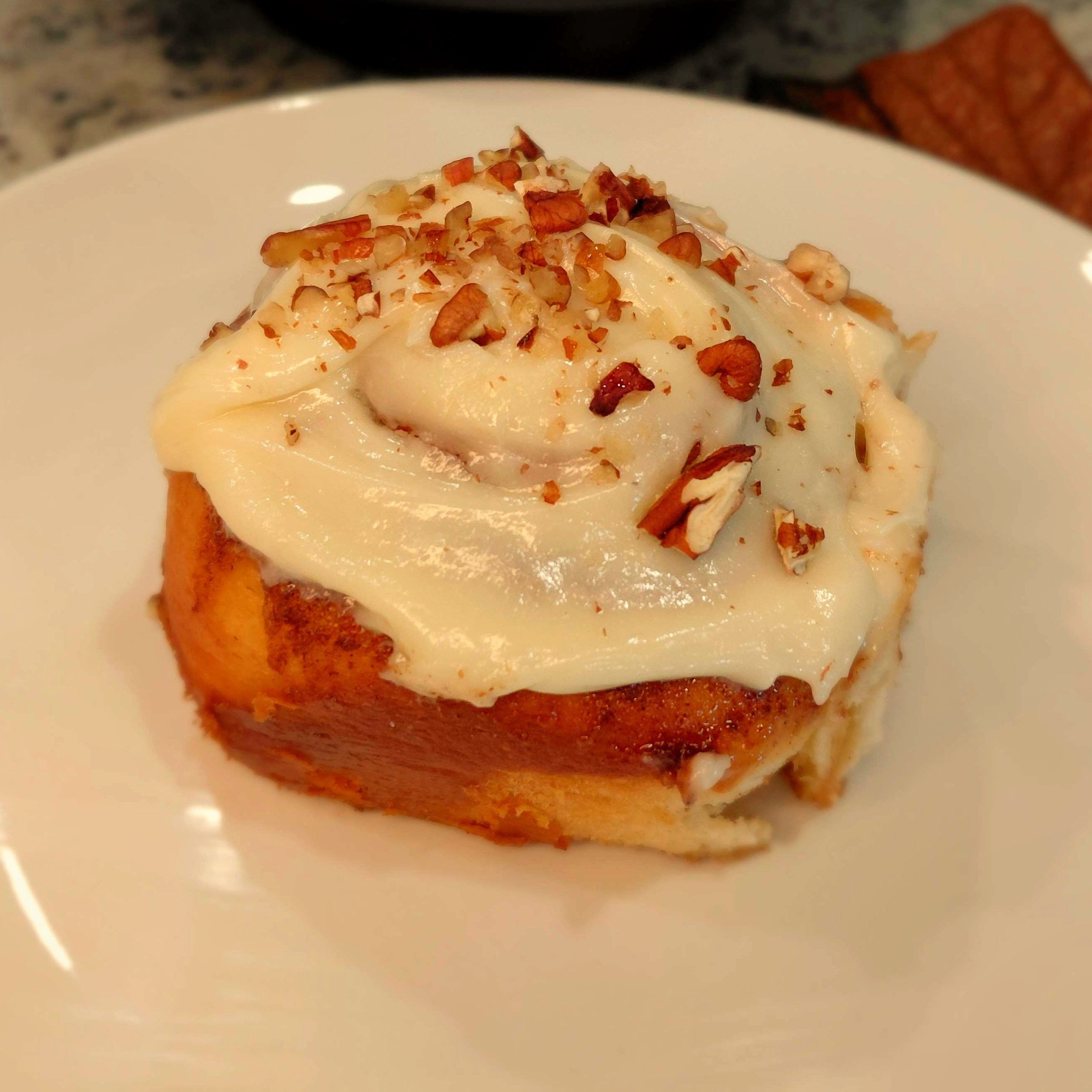 Cinnamon roll with cream cheese frosting and chopped pecans on a white plate