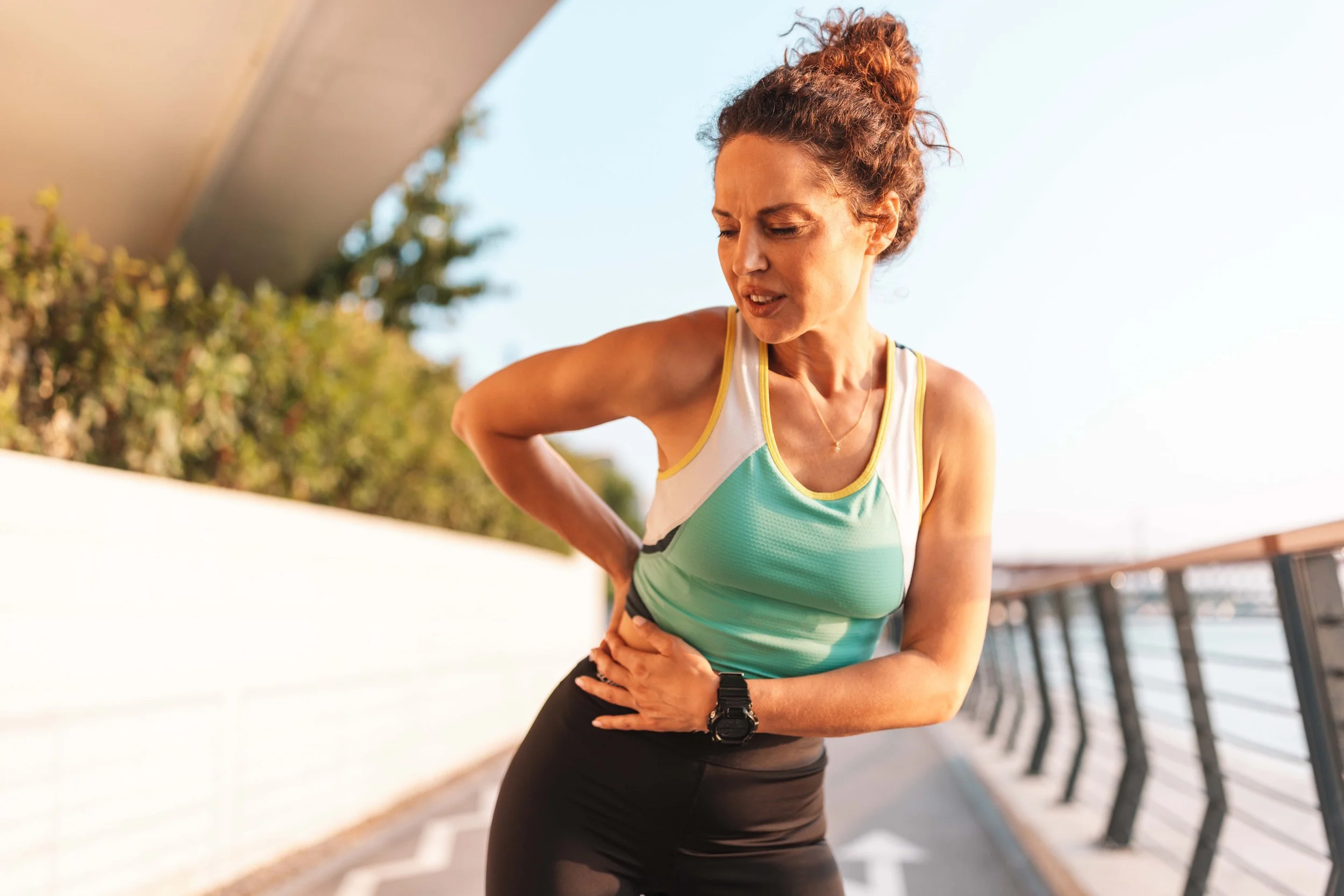 A woman in athletic wear, gripping her side in pain, outdoors on a sunny day, near a row of benches.