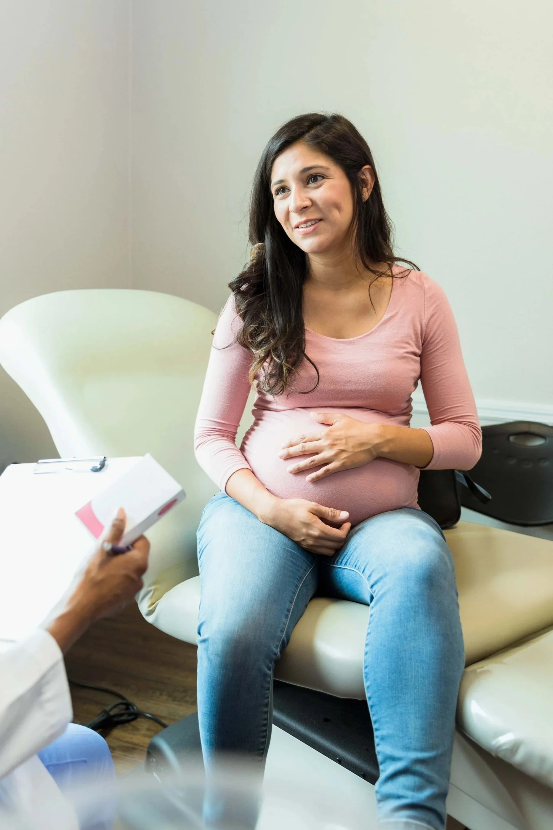 Pregnant woman sitting on a medical examination table during a prenatal checkup, with her hand on her belly, in a clinic setting.
