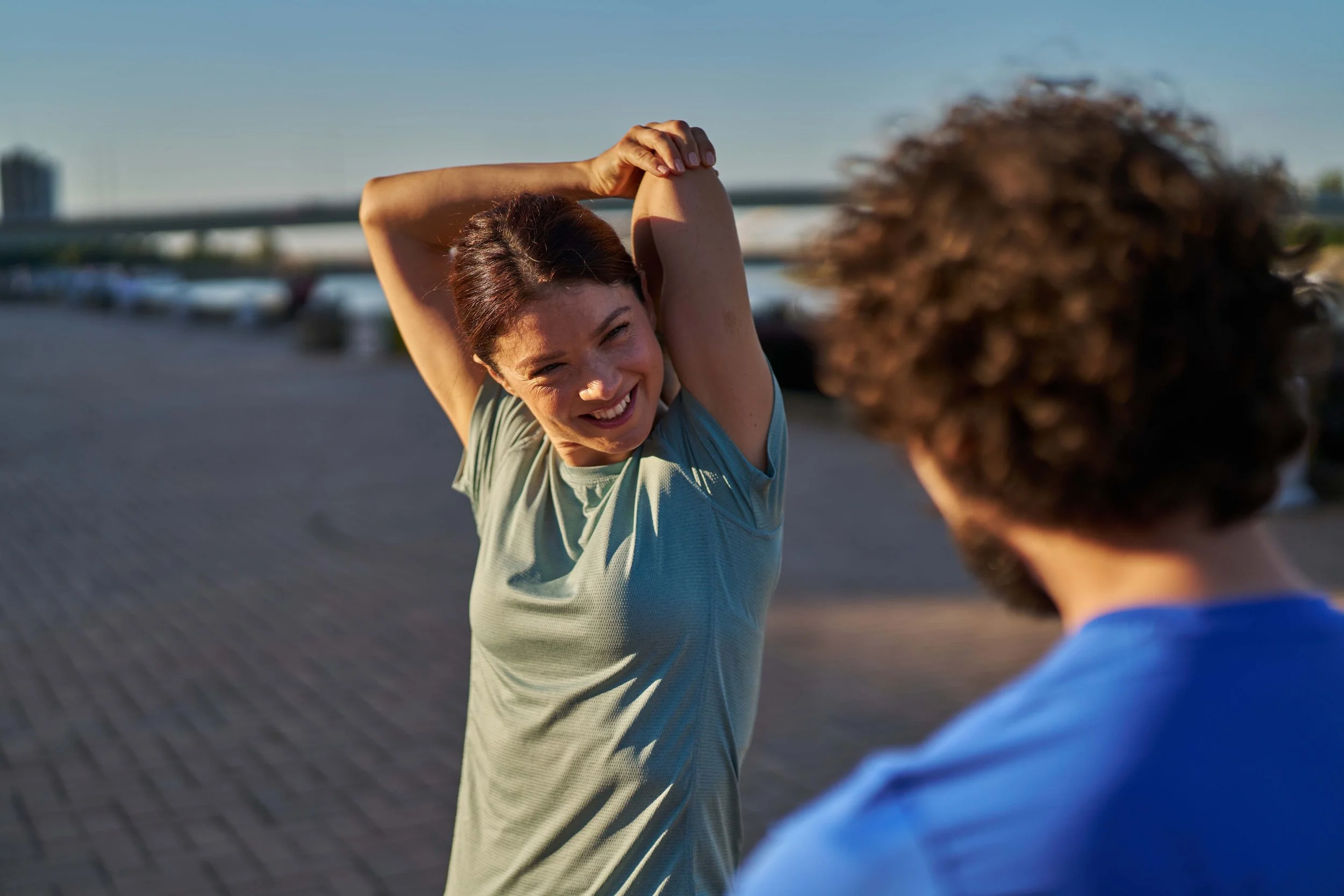 A woman with short dark hair smiling and stretching arms behind her head outdoors, with a blurred person in the foreground and a bridge visible in the background on a clear day.