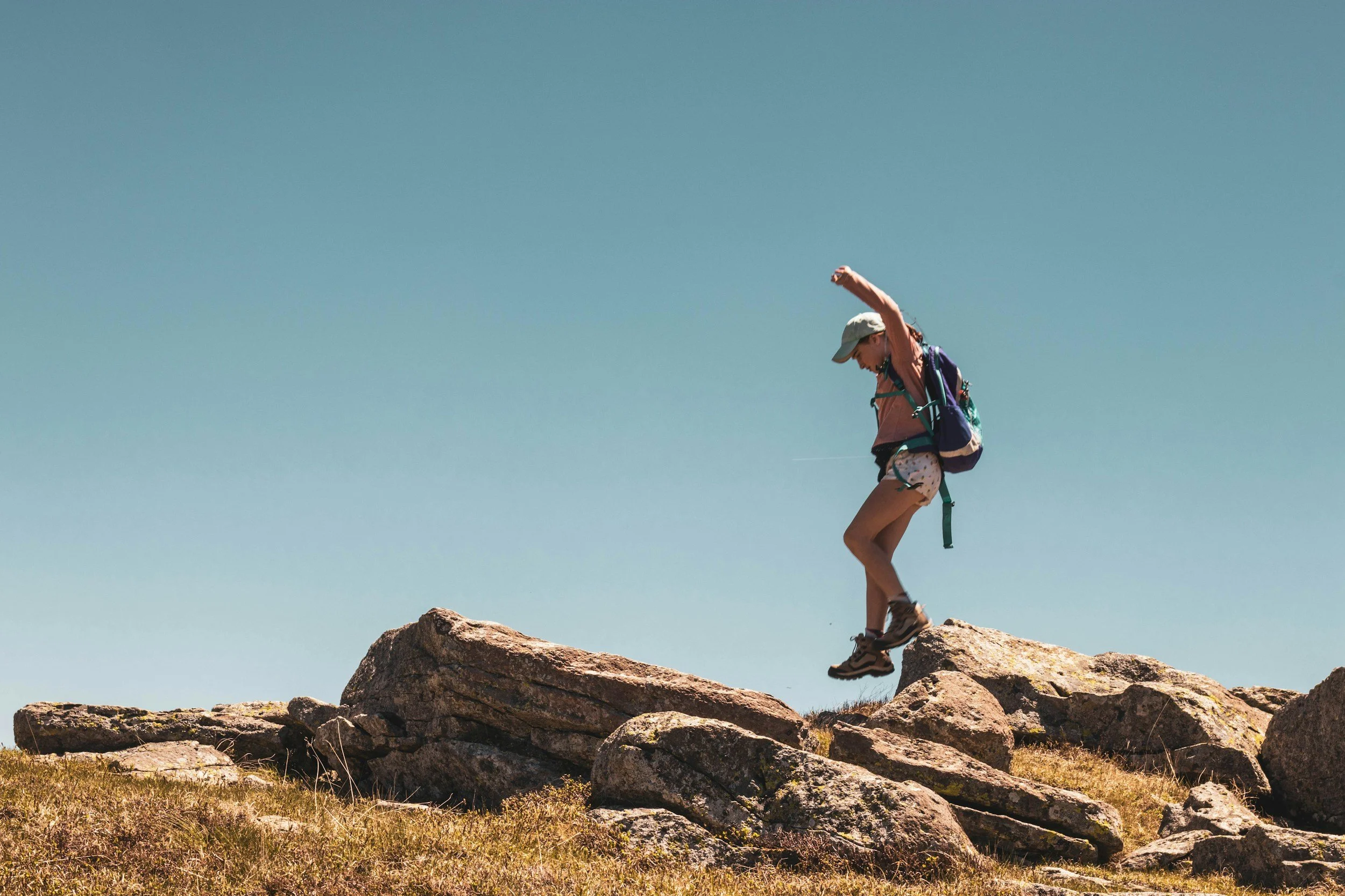 A person standing on large rocks outdoors under a clear blue sky, wearing a hat, backpack, and hiking shoes, with one arm raised.