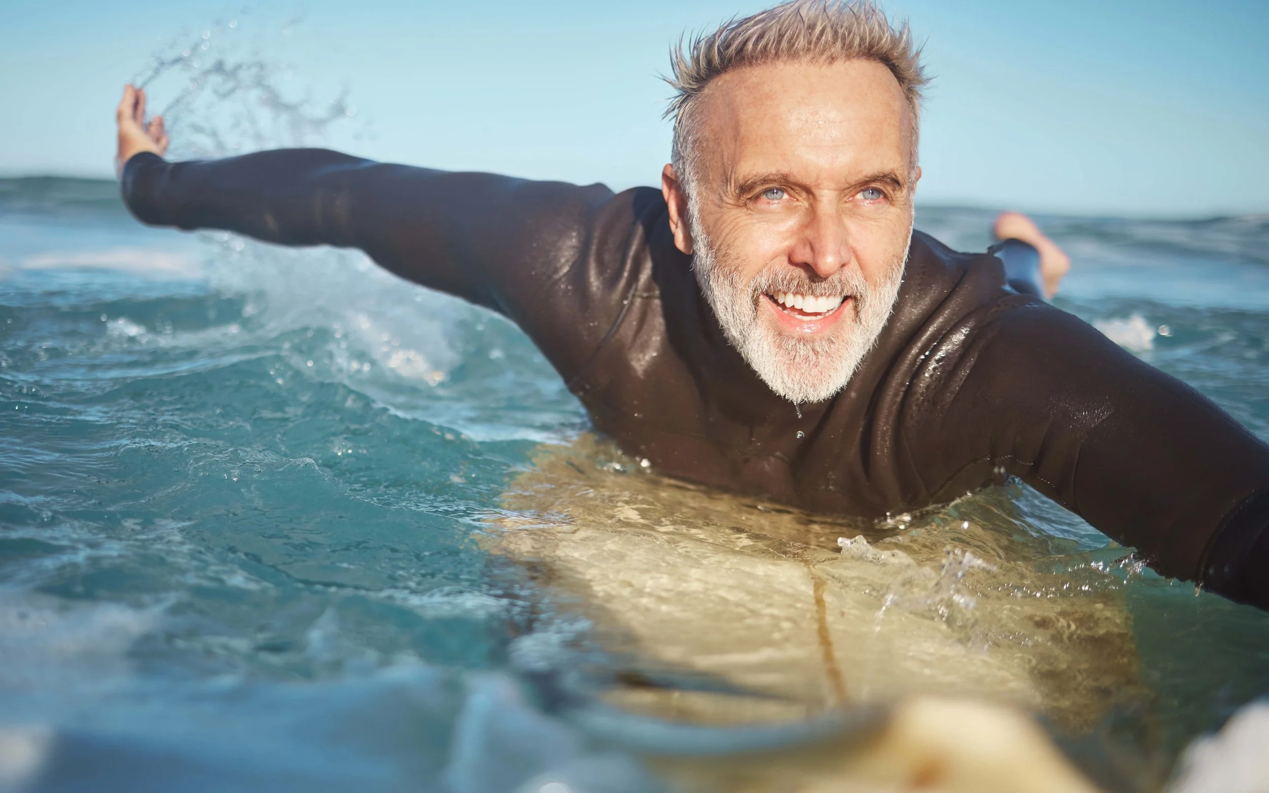 A smiling elderly man with gray beard and hair, lying on a surfboard in the ocean, wearing a black wetsuit, with arms extended to balance in the water.