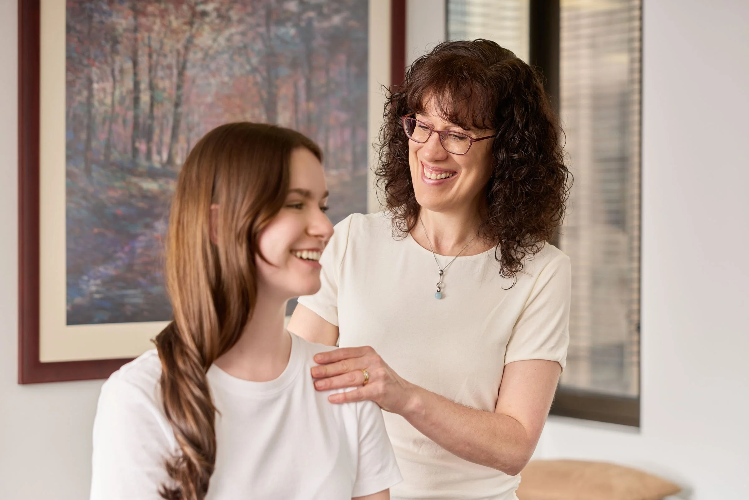 A woman with curly brown hair and glasses smiling at a young girl with straight brown hair, who is smiling as well, inside a room with a landscape painting in the background.