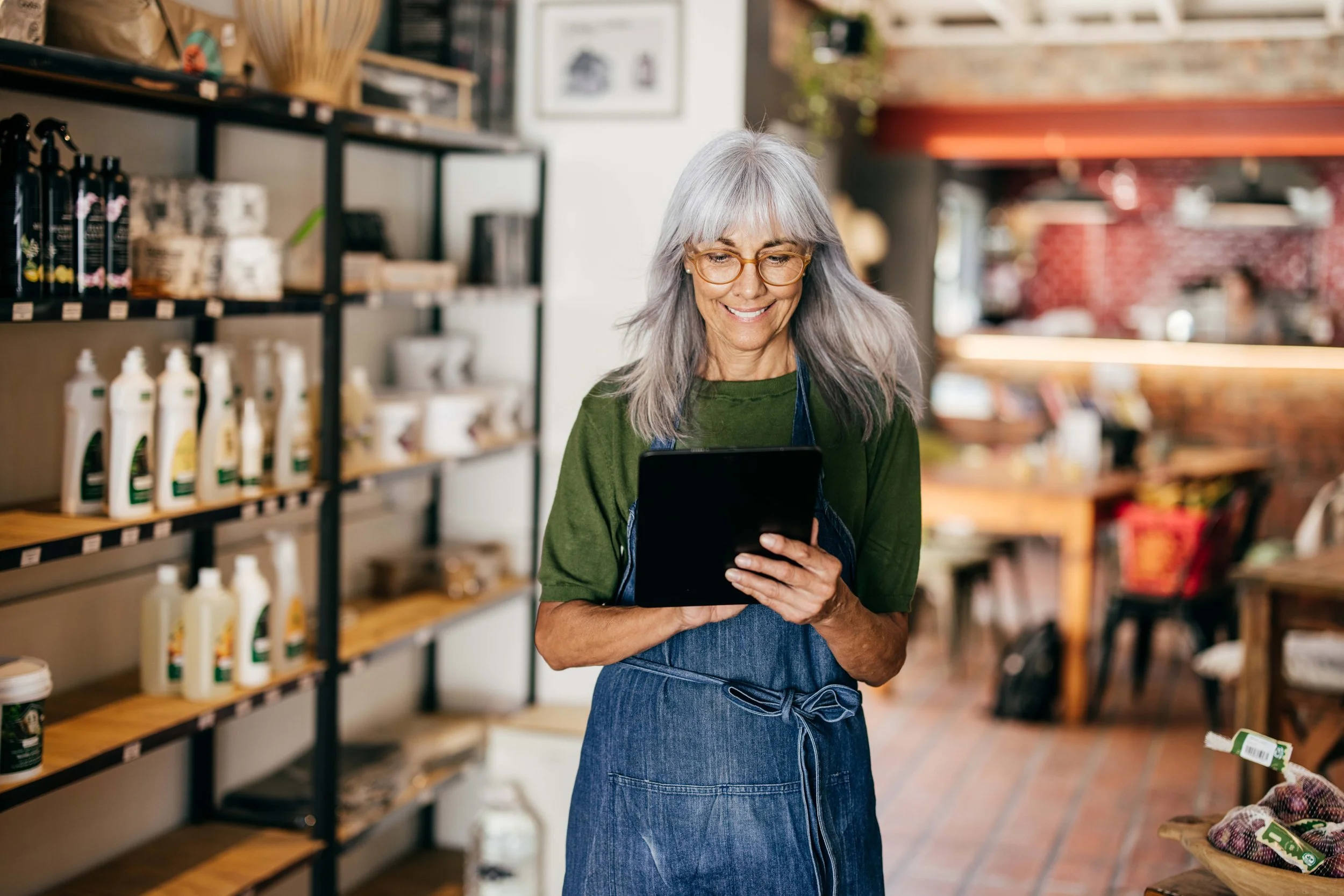 A smiling elderly woman with gray hair and glasses, wearing a green shirt and denim apron, shopping in a grocery store, looking at a tablet device.
